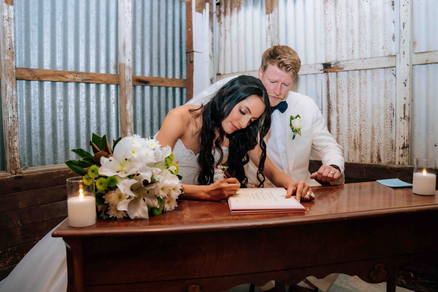 Bride and groom signing wedding register at rustic wooden table with white floral bouquet and candles.