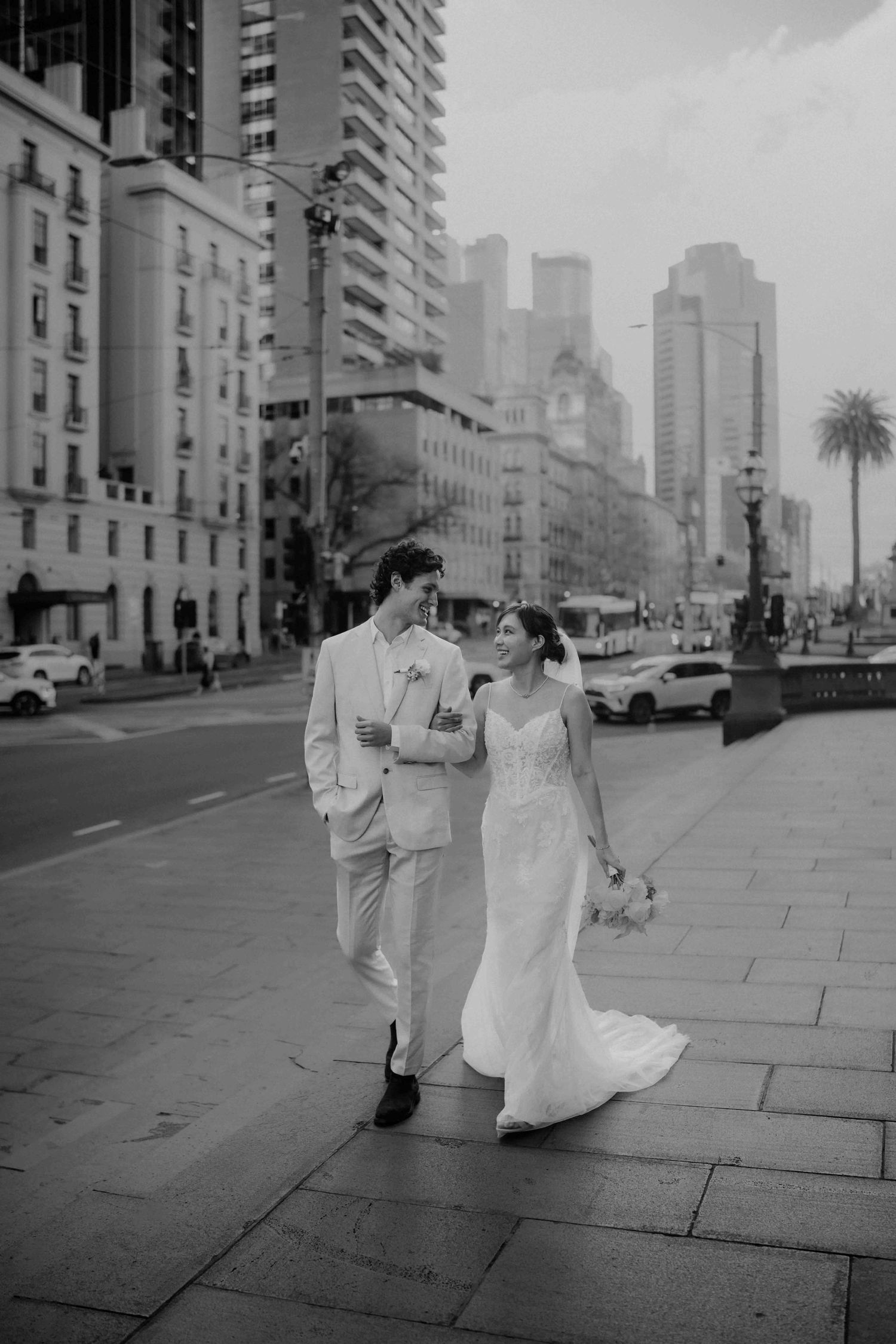 Bride and groom walking together on city sidewalk with urban skyline in black and white.