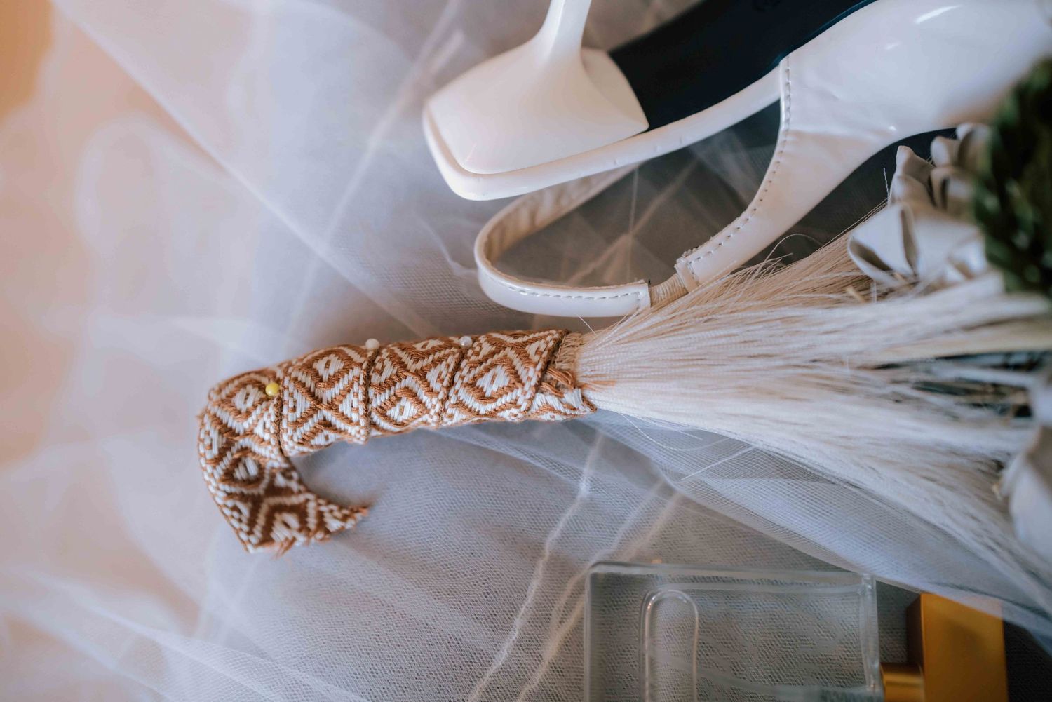 Decorative patterned bouquet handle resting on white tulle veil beside bridal heels.