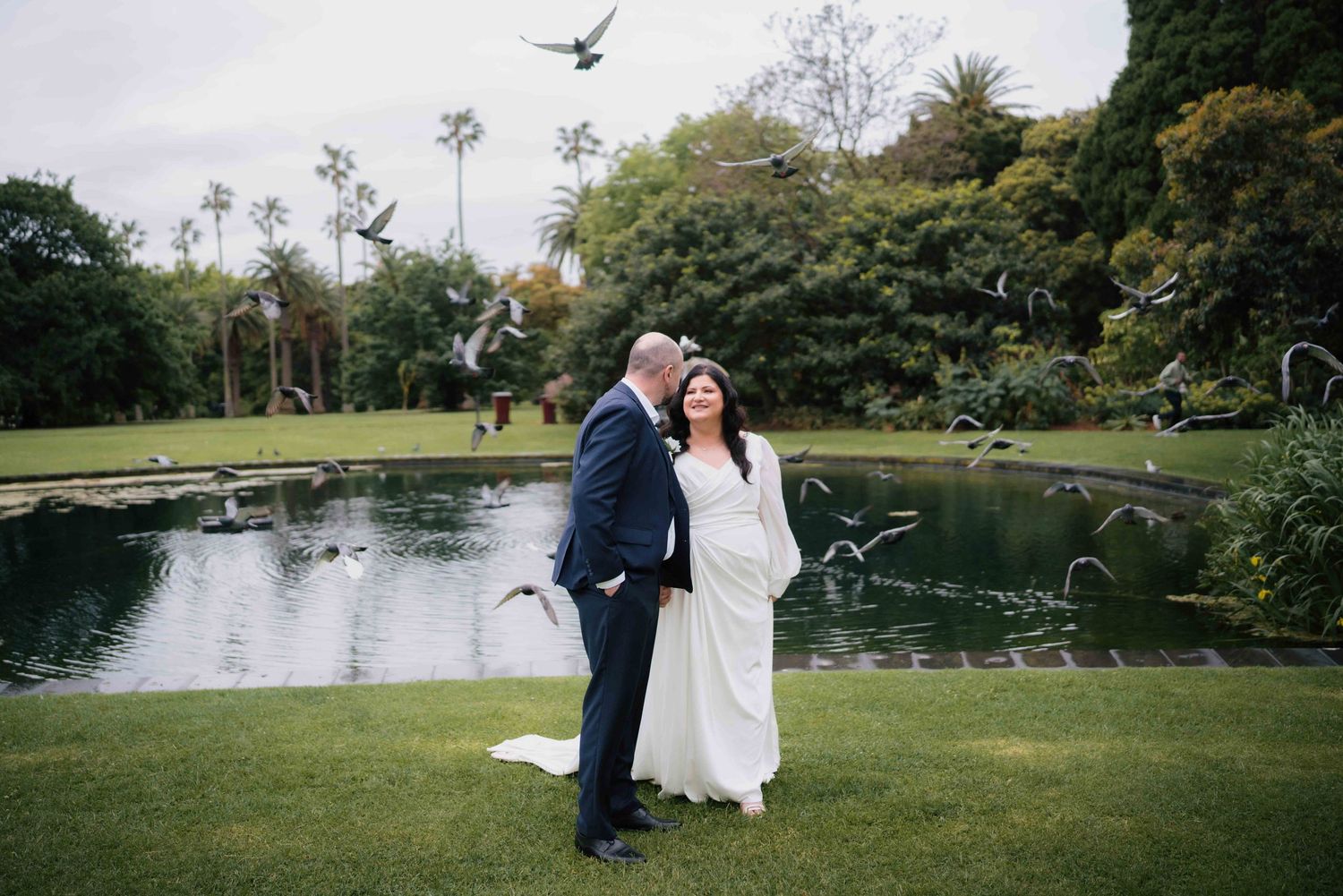Bride and groom embrace in a scenic garden with birds in flight above a tranquil pond.