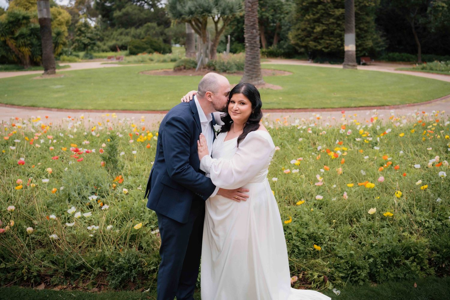 Groom kissing bride on the cheek in front of a colorful wildflower garden.
