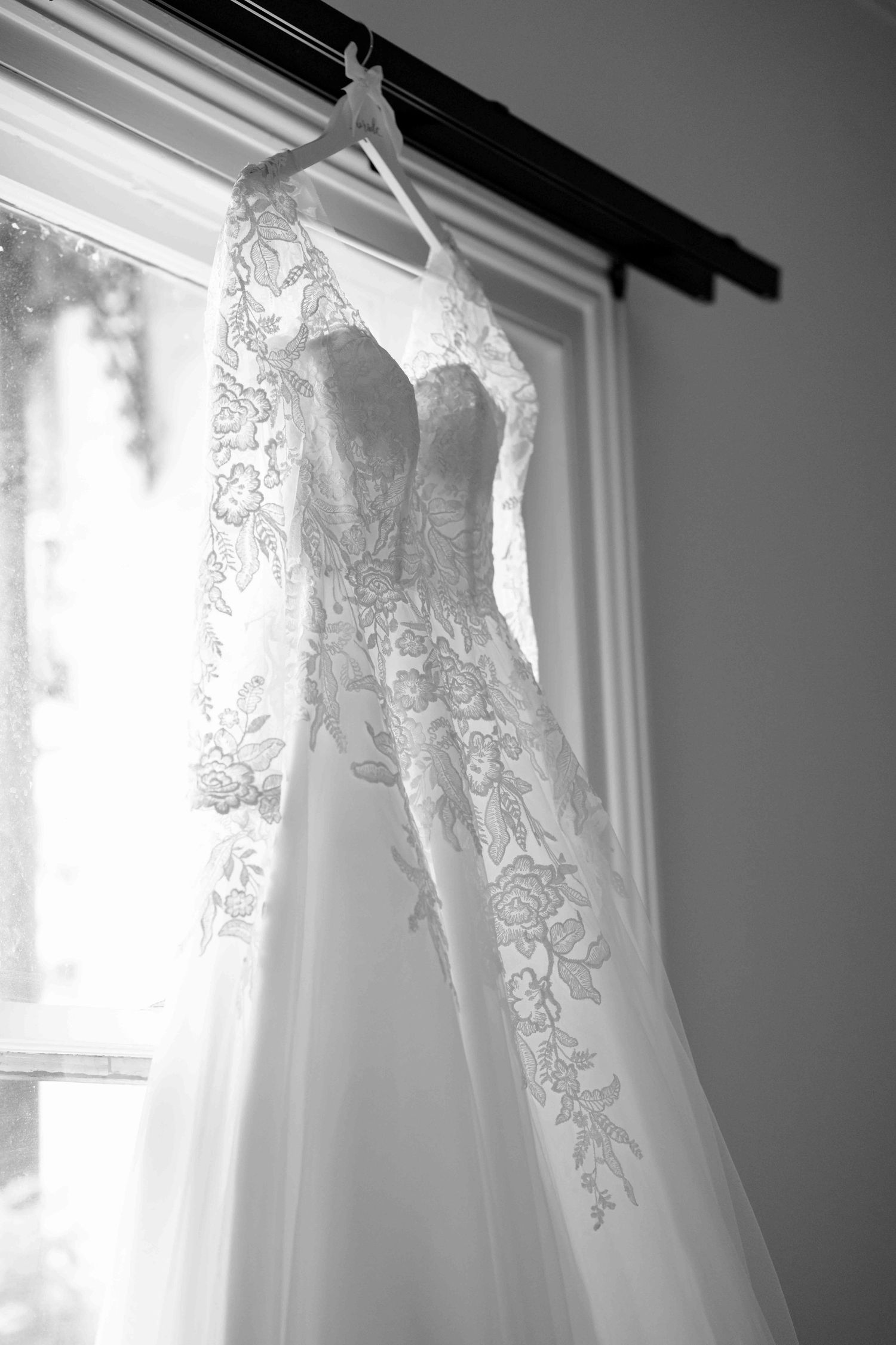 Beautiful lace wedding dress hanging by window on wooden hanger before ceremony.