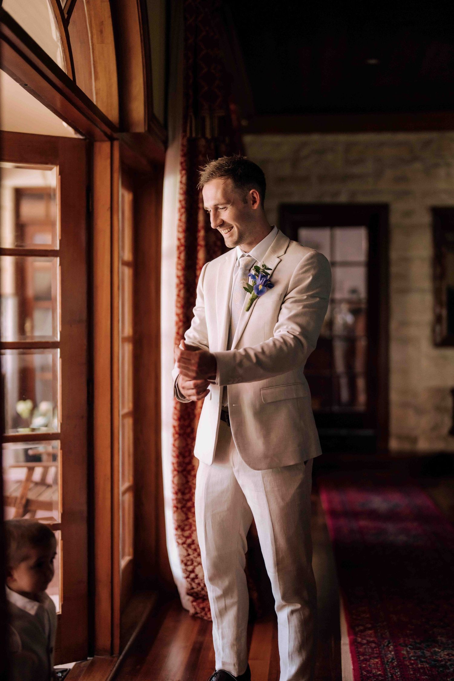 Smiling groom in cream suit with floral boutonniere stands by wooden door in warm light.