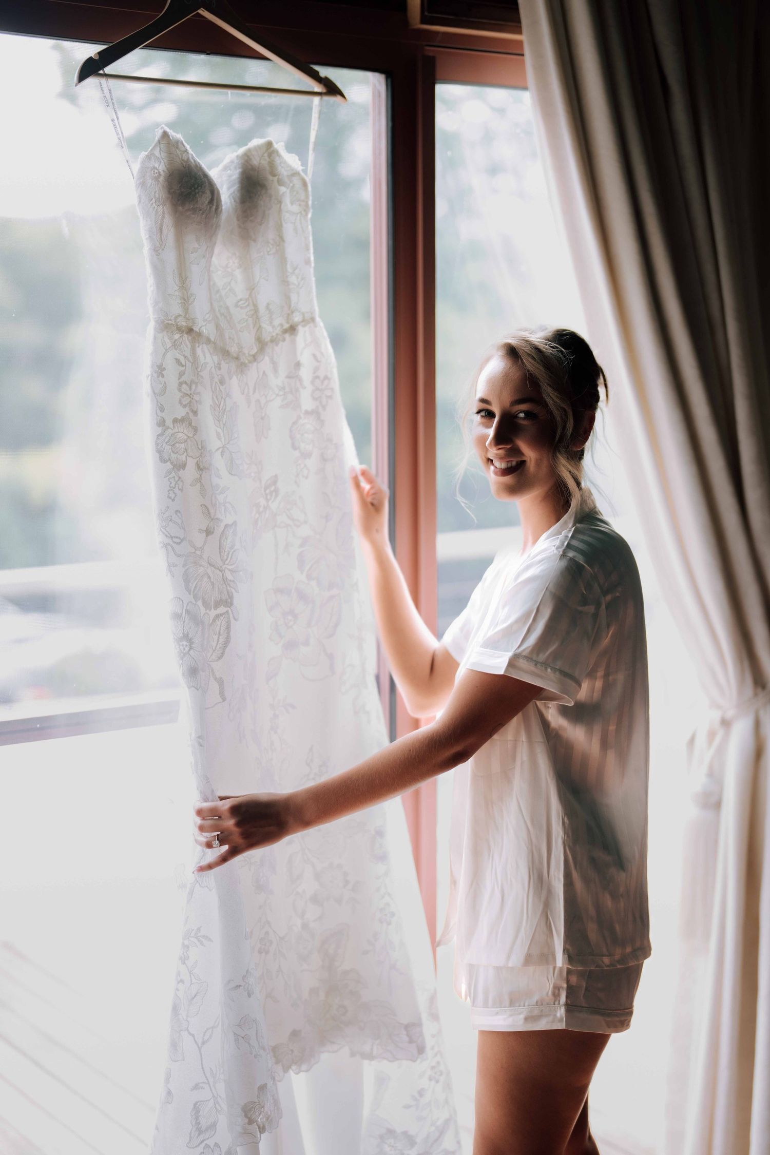 Smiling bride in white pajamas standing beside her lace wedding dress hanging by a sunlit window.
