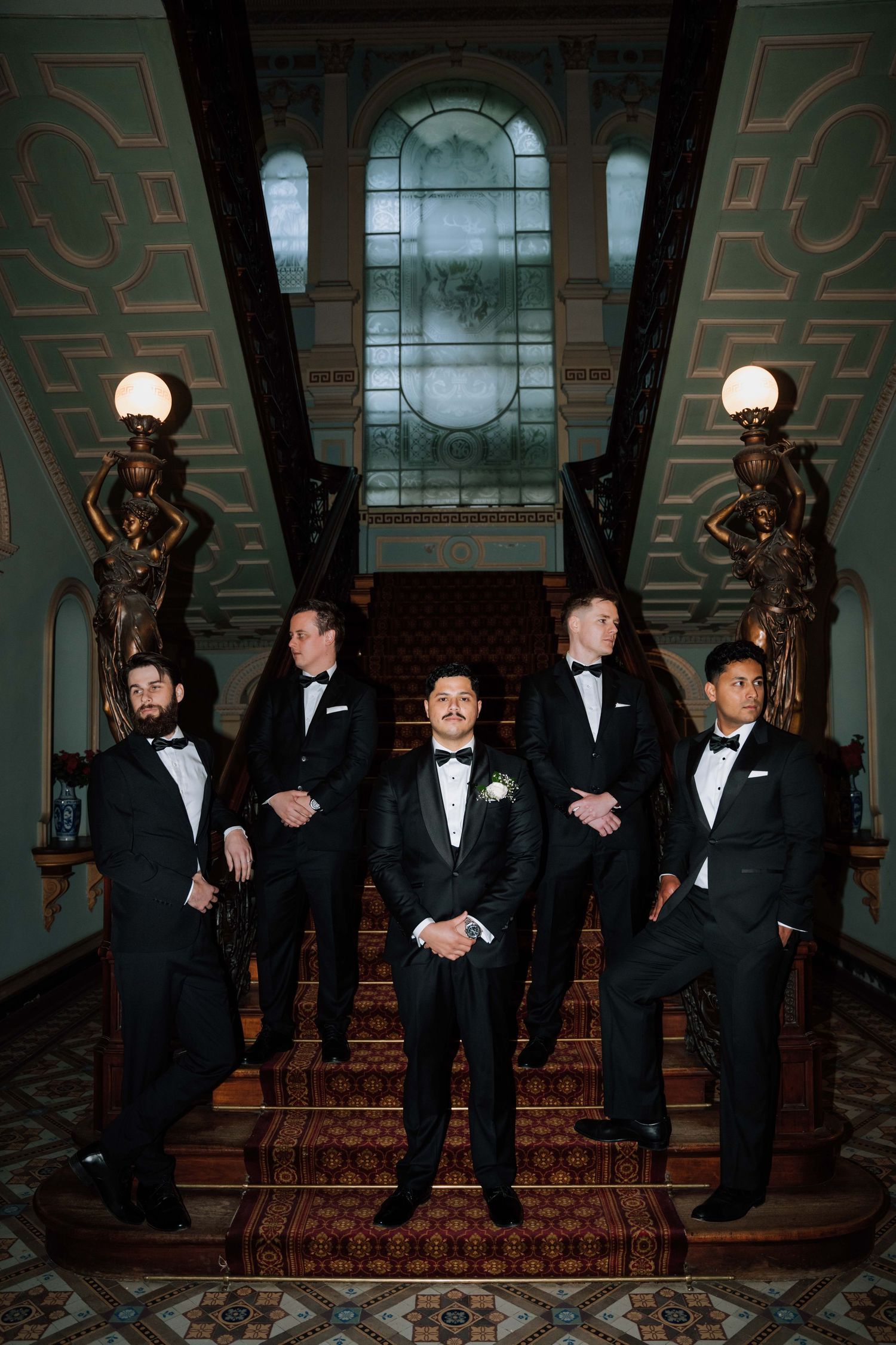 Groom and four groomsmen in black tuxedos posing on grand staircase with stained glass window backdrop.