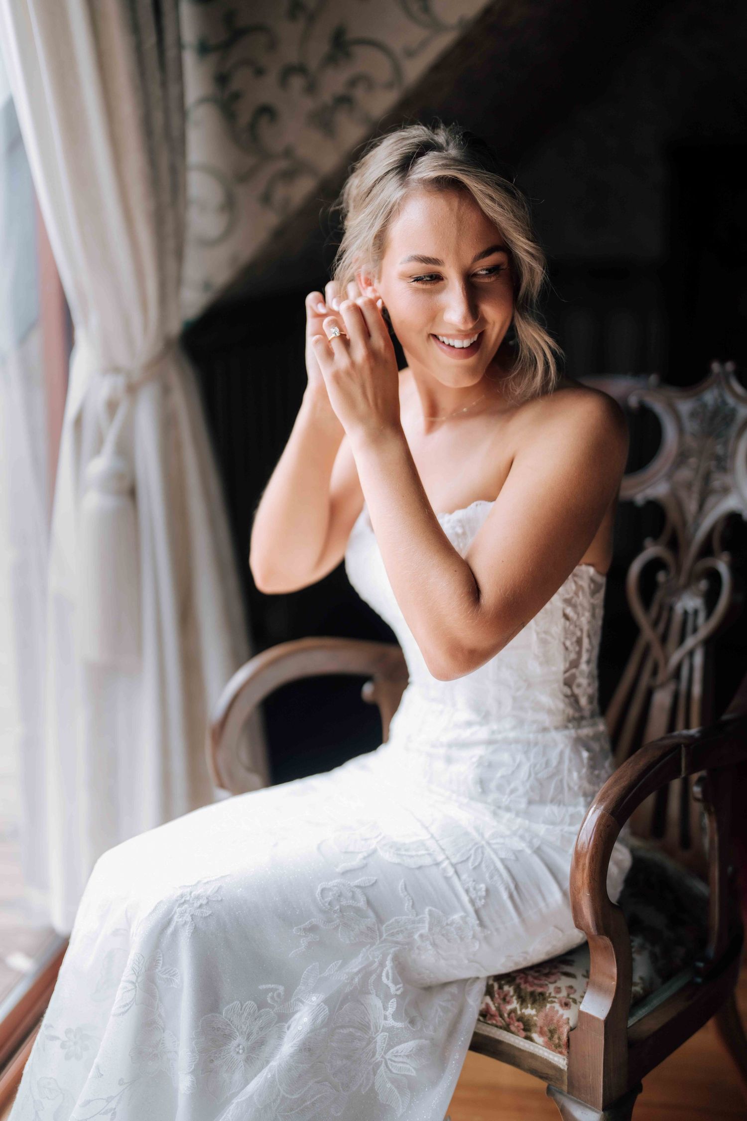Smiling blonde bride in strapless lace wedding gown puts on earrings while seated in vintage chair.