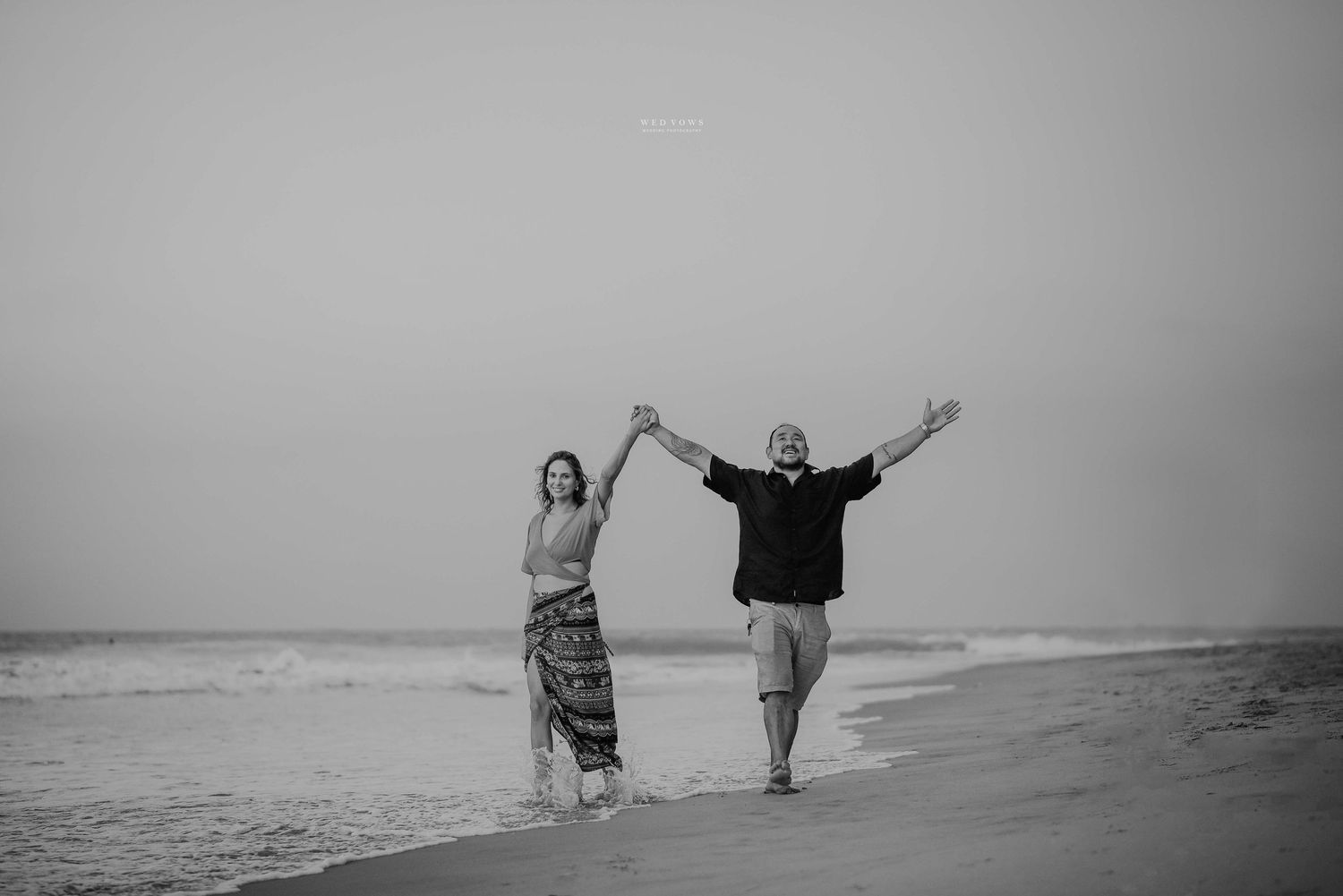 Black and white photo of joyful couple splashing in ocean waves on sandy beach.