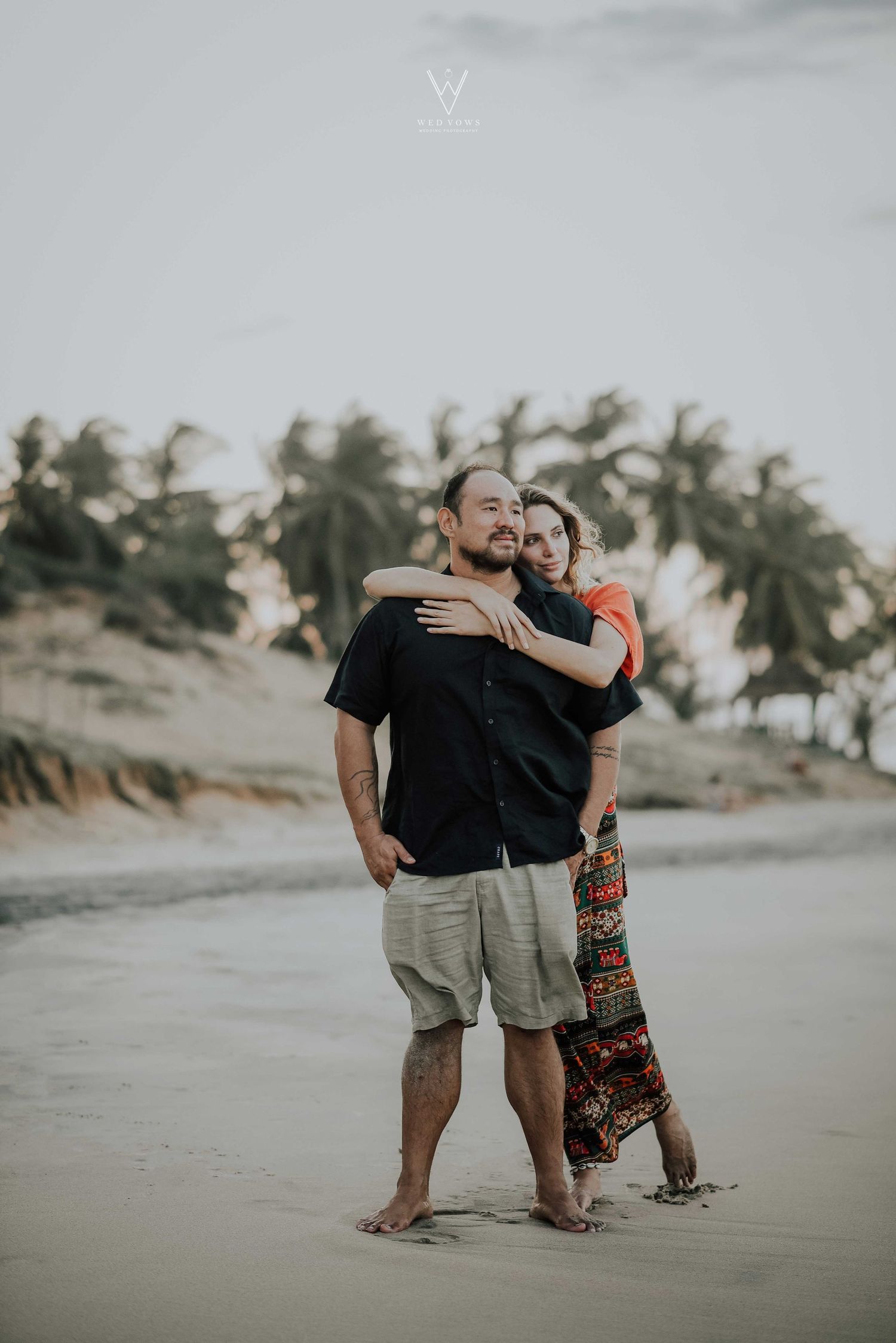 Woman hugging man from behind while standing on a tropical beach with palm trees.