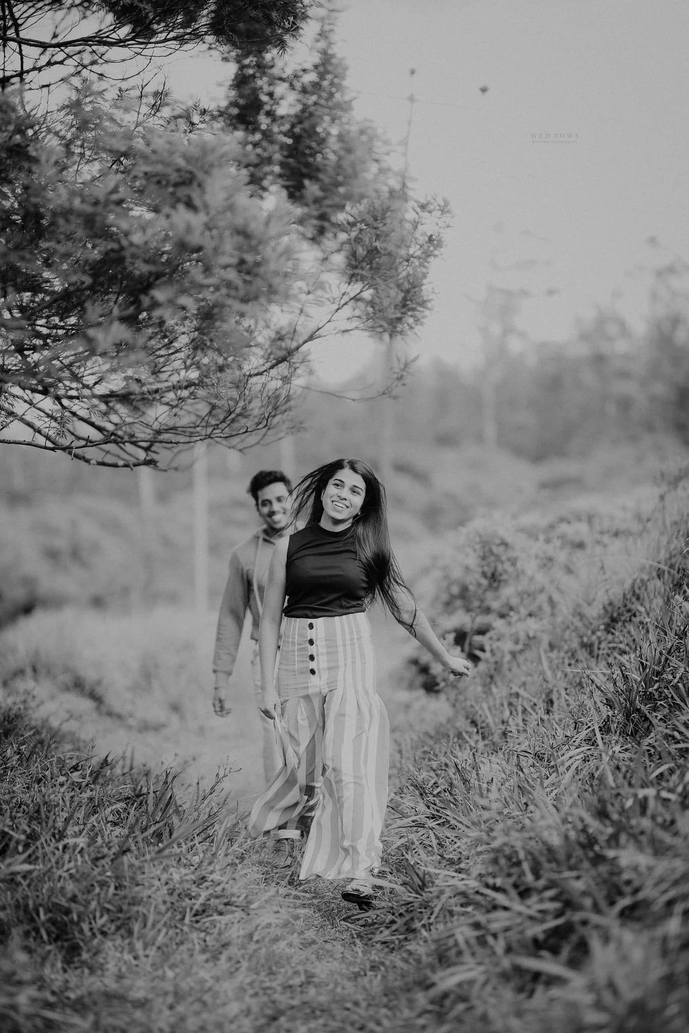 Black and white photo of couple walking on a forest trail with woman leading and smiling at camera.