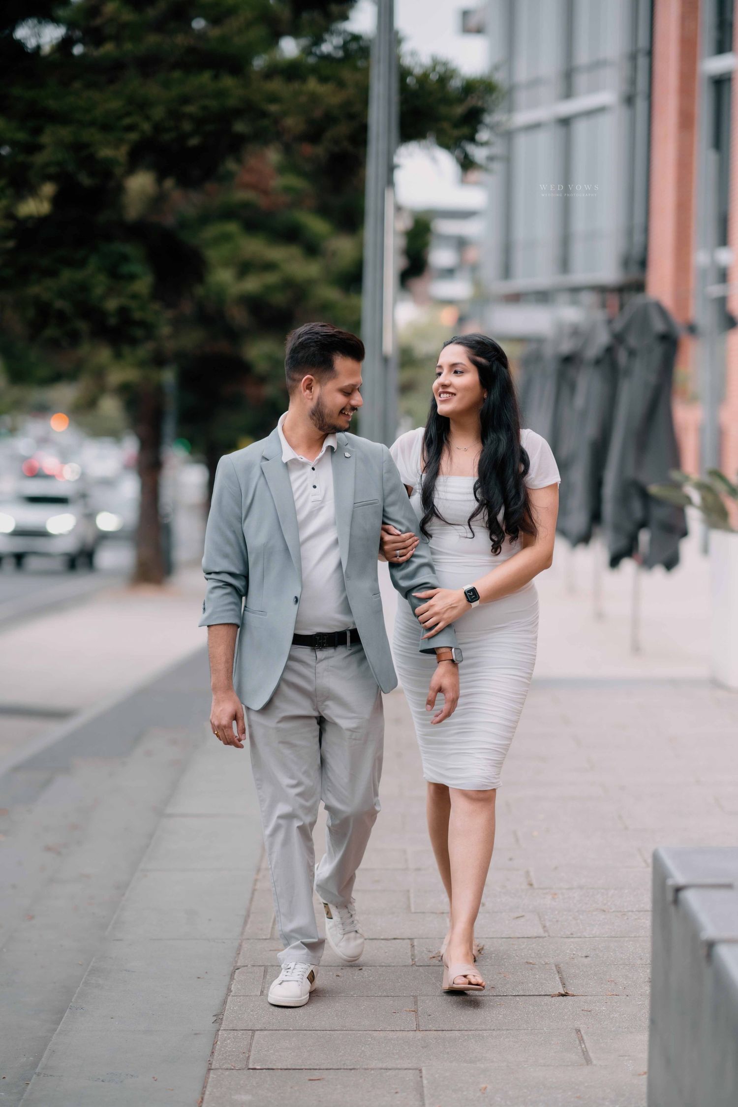 Smiling couple in matching grey and white outfits walk together along a city sidewalk.
