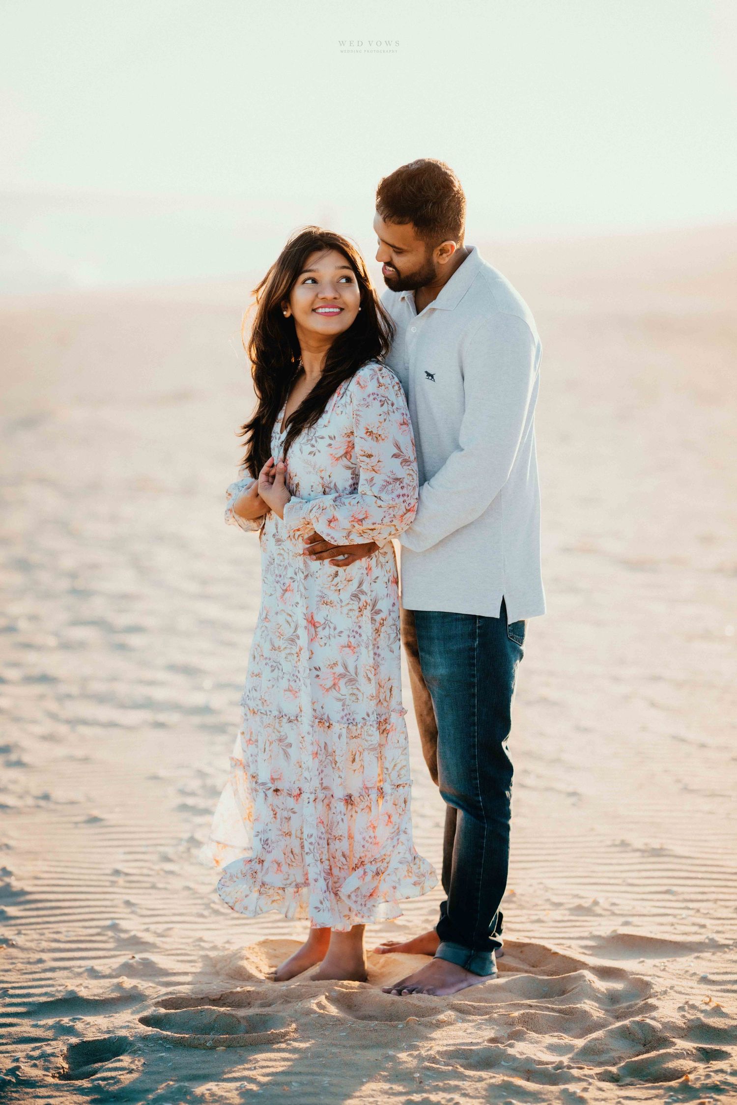 Couple embracing barefoot on sandy beach dunes, woman in floral dress smiling up at partner in golden light.