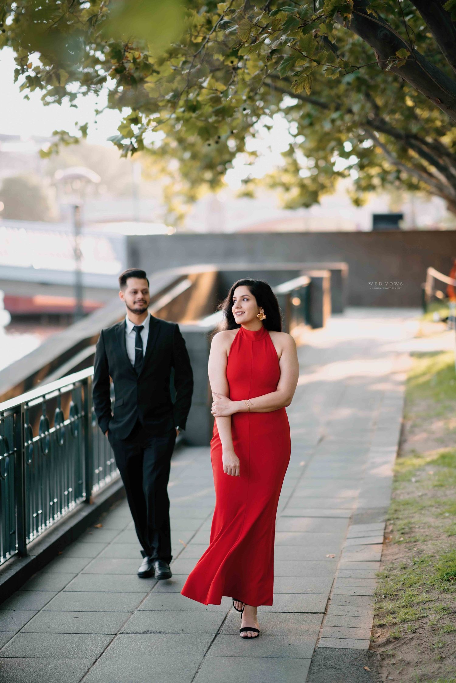 Woman in red halter maxi dress walks confidently along a riverside path with man in black suit.