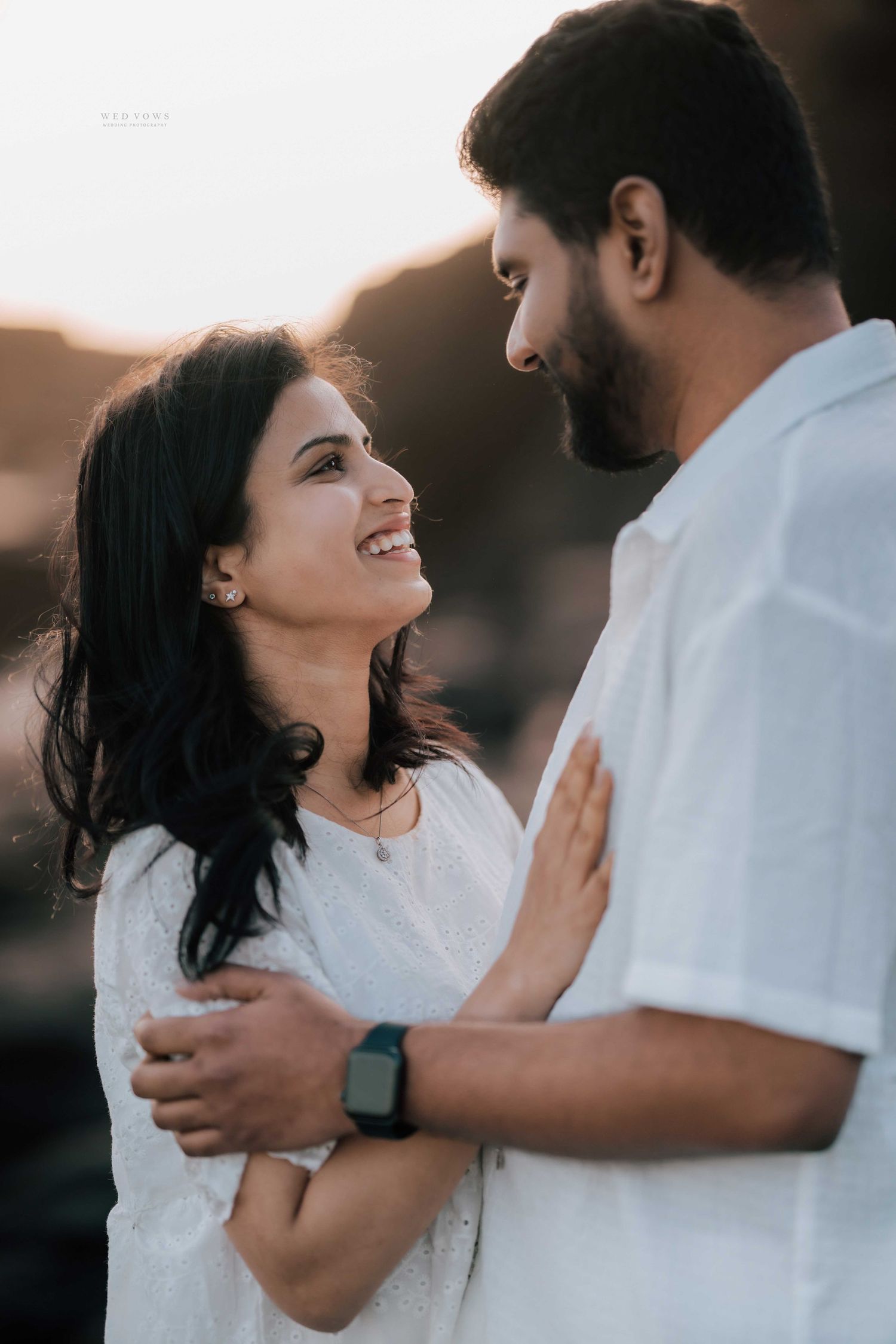 Joyful woman smiling up at bearded man during romantic golden hour coastal photo shoot.