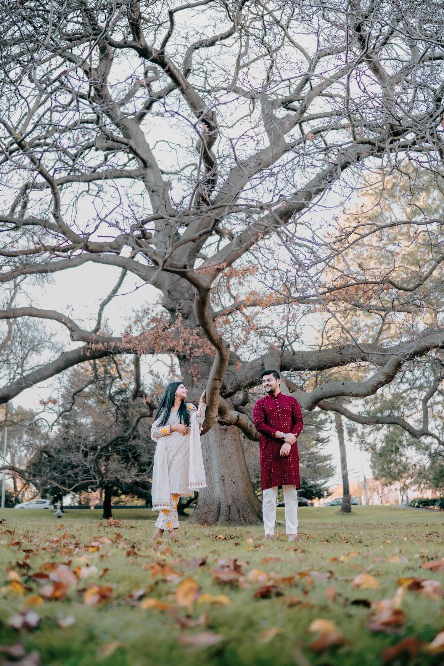 Couple in traditional Indian attire standing apart near a massive bare tree trunk in an autumn park.