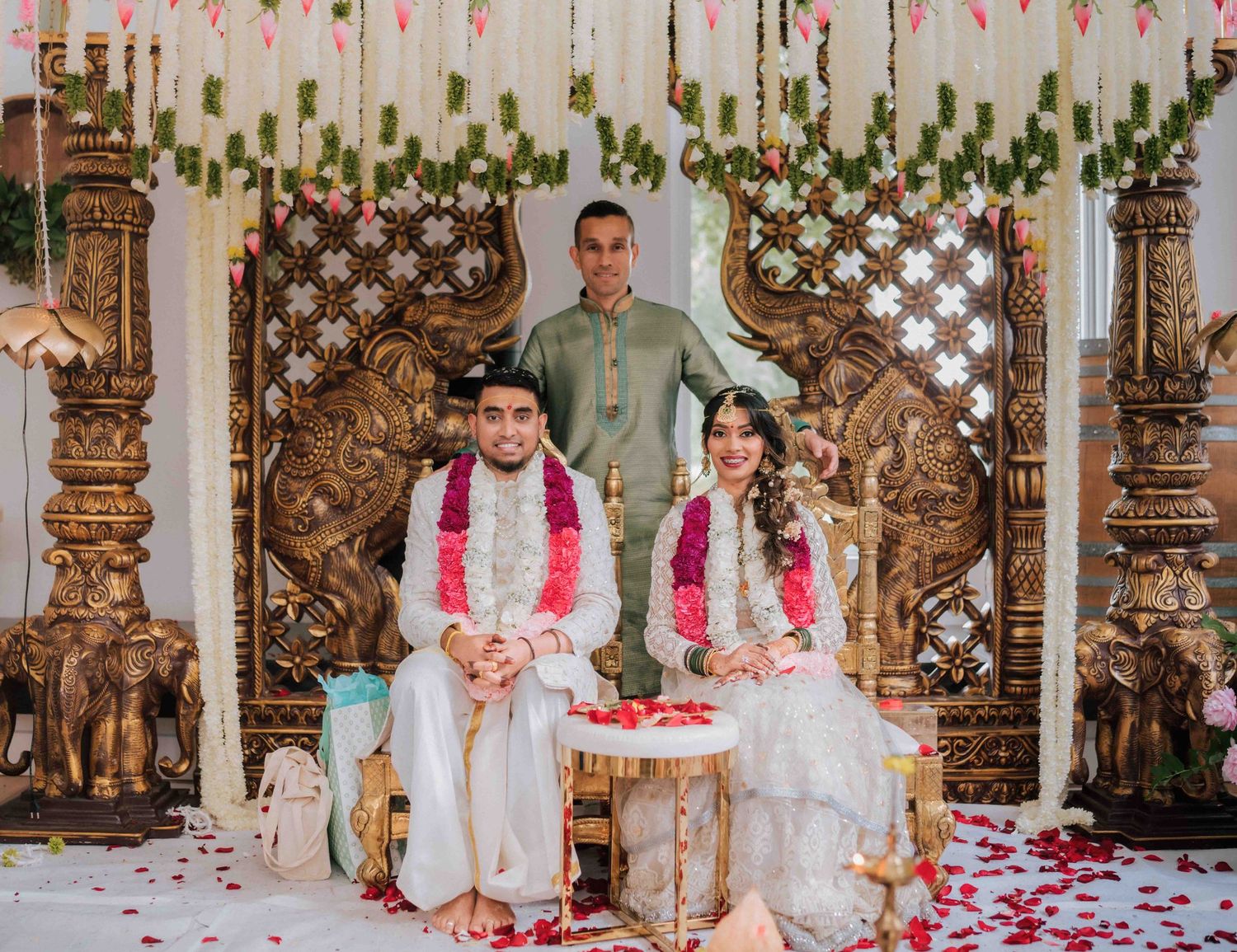Bride and groom posing at carved golden mandap with rose petals scattered on white floor.