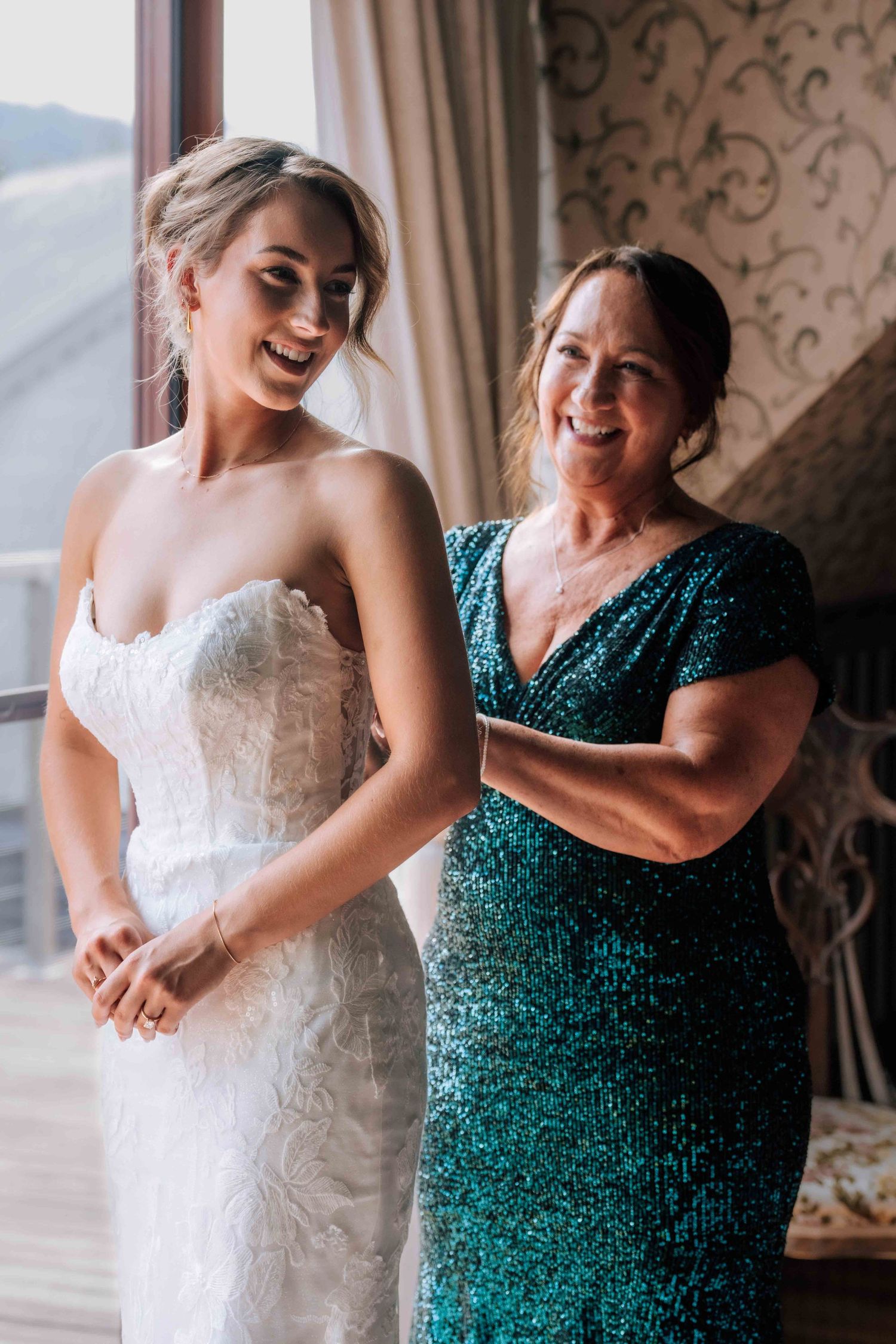 A bride in a white lace gown laughs joyfully as a woman in a teal sequin dress helps her prepare for her wedding.