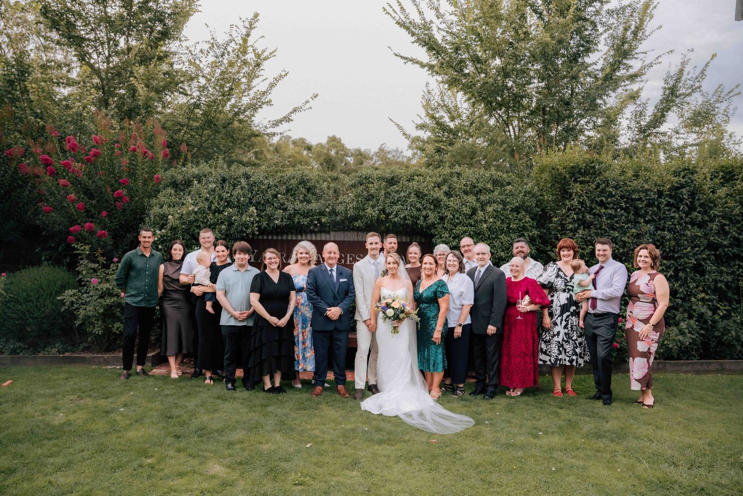 Wedding party group photo with bride and groom surrounded by family on a green lawn with lush hedges.