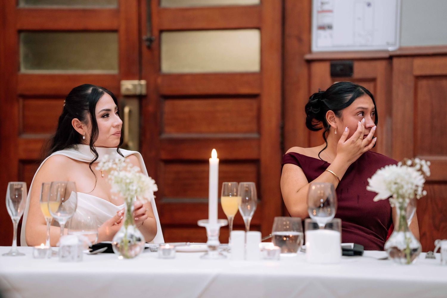 Bride and bridesmaid seated at wedding reception table, bridesmaid covering mouth emotionally during heartfelt speeches.
