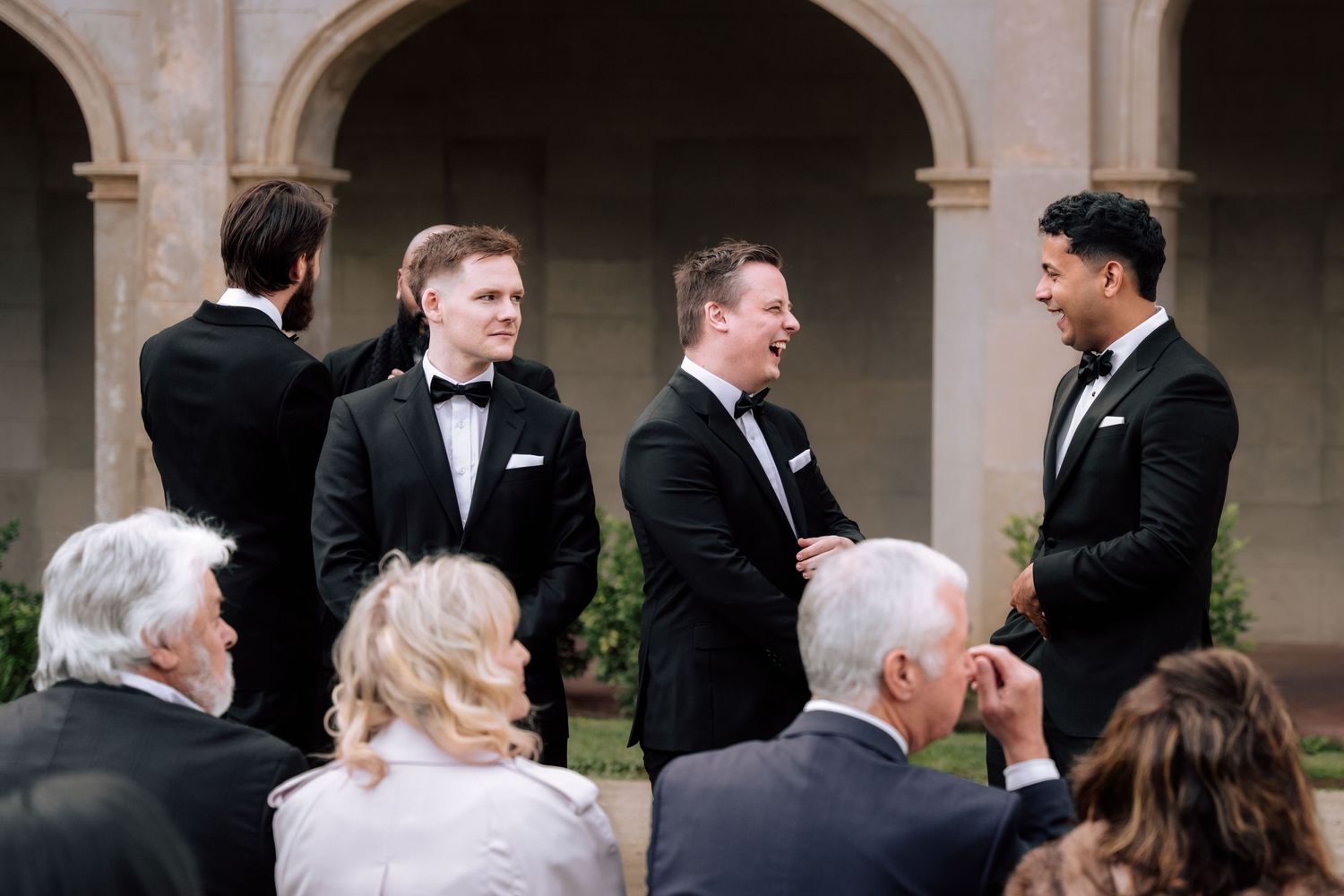Groomsmen in black tuxedos socializing with wedding guests before the outdoor ceremony begins.