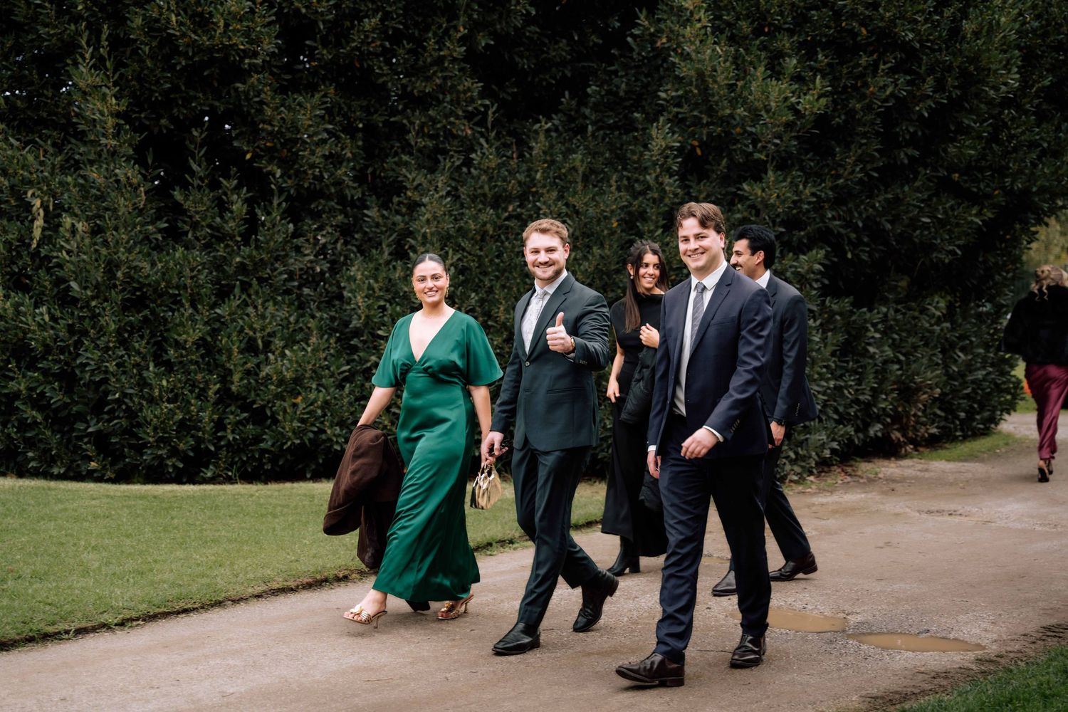 Wedding guests in formal attire walking along garden path toward ceremony venue.