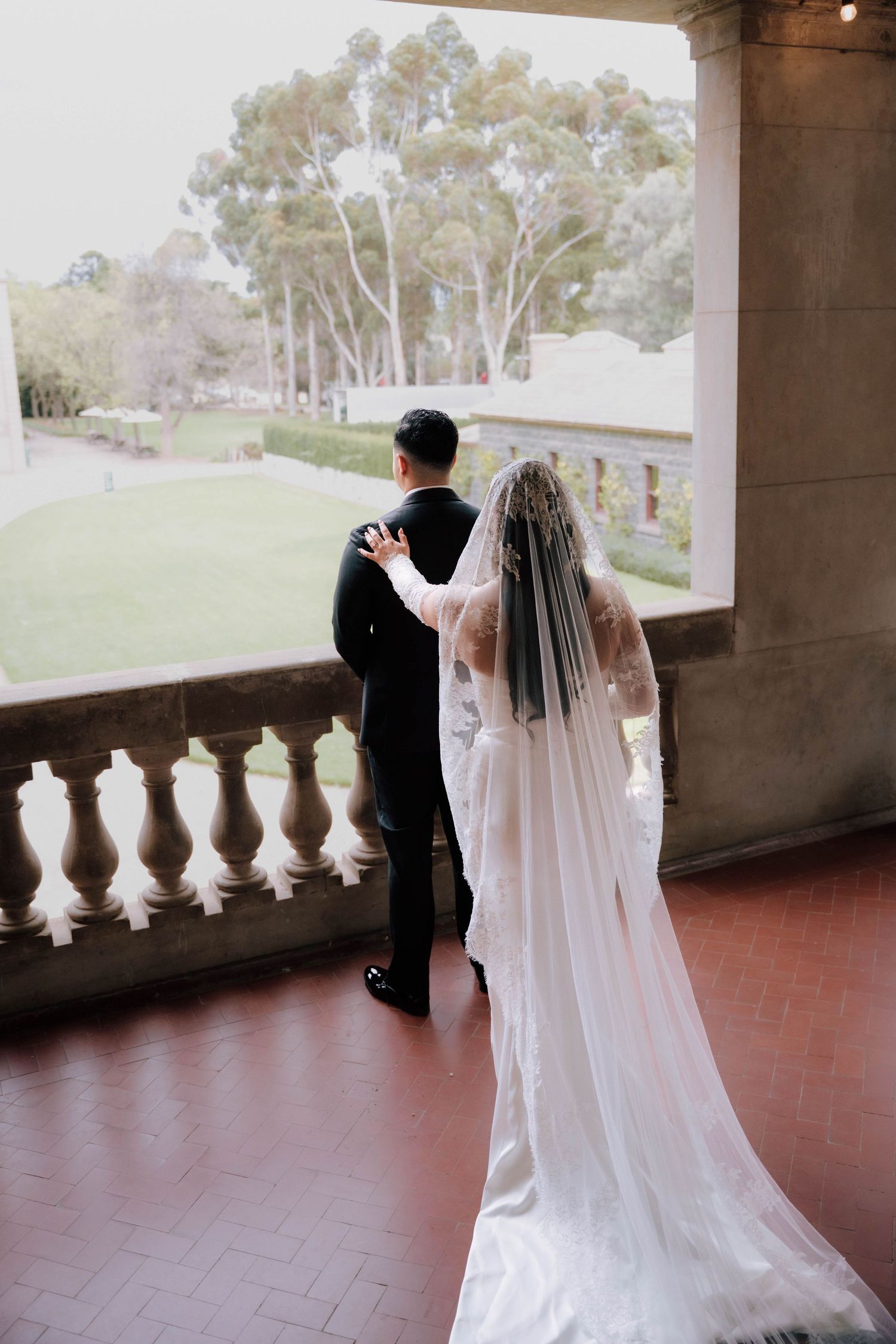 Bride in white gown and veil stands with groom in black tuxedo on ornate balcony overlooking lush garden.