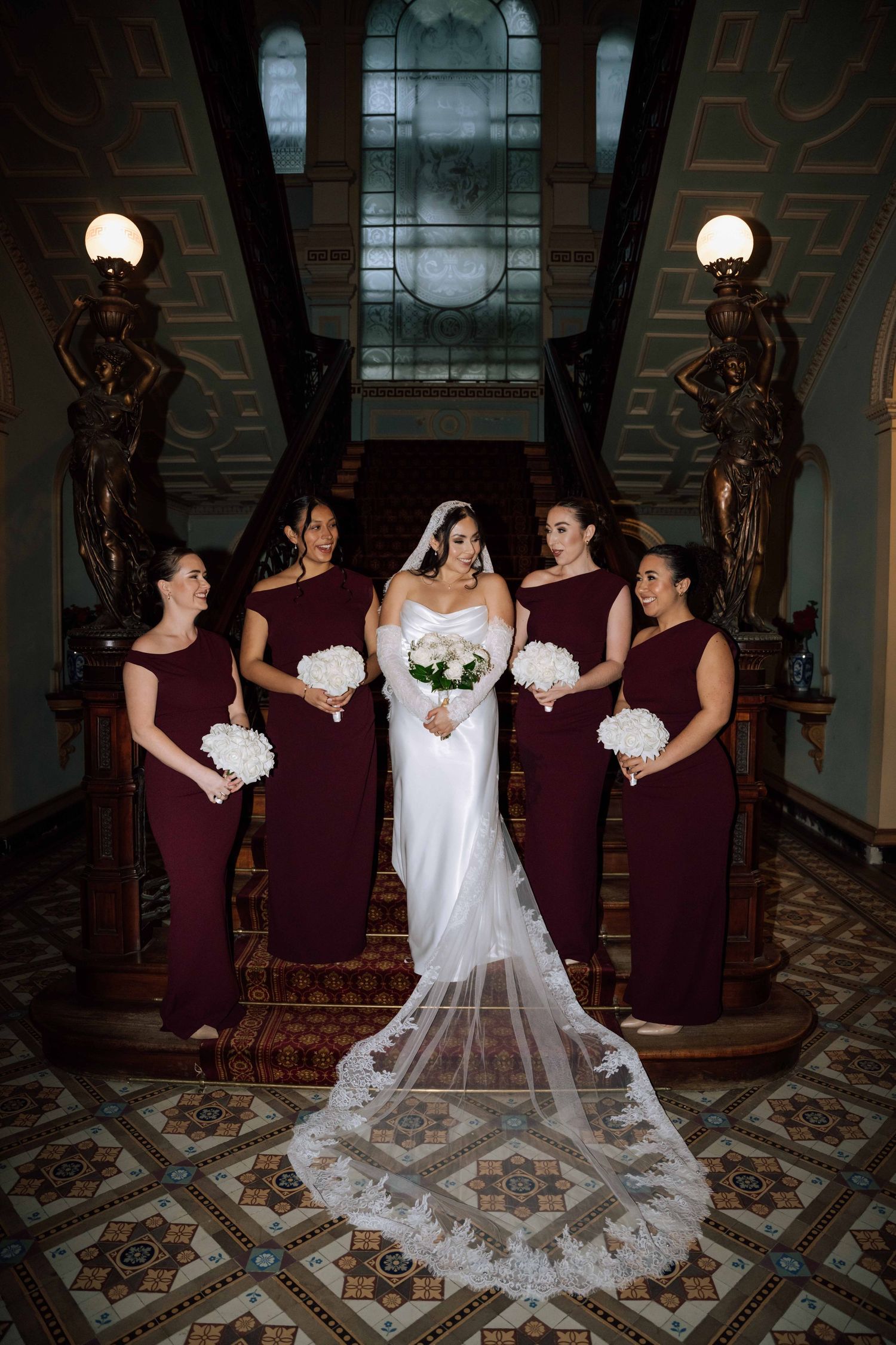 Bride in white gown with bridesmaids in burgundy dresses on ornate staircase.