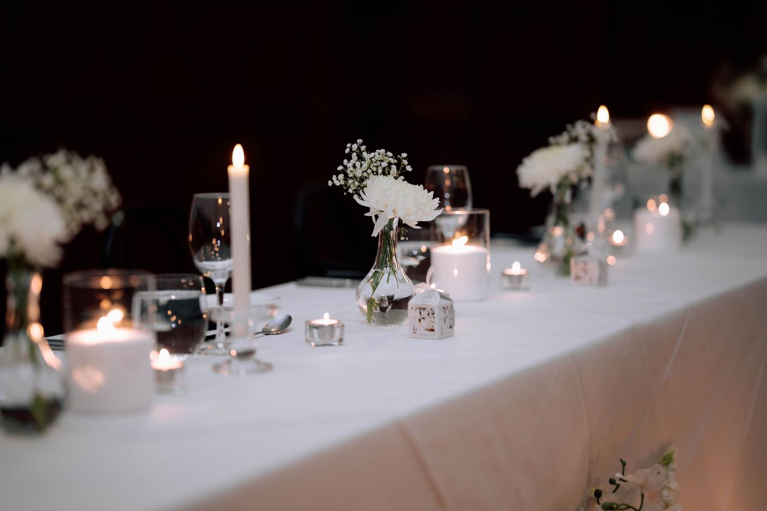 Elegant wedding reception table with white flowers, candles, and glassware in soft candlelight.