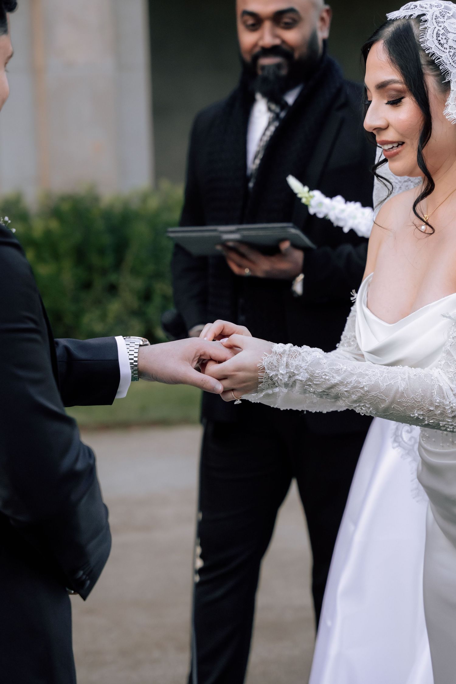 Bride and groom exchanging rings during outdoor wedding ceremony with officiant holding book behind them.