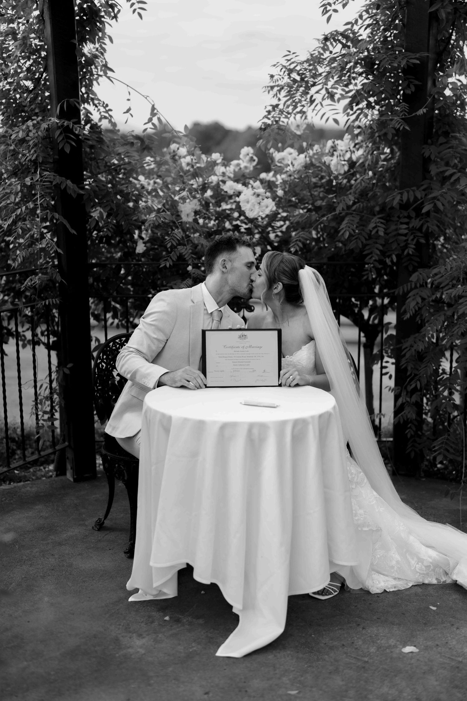 Black and white photo of bride and groom sharing a kiss at signing table during wedding ceremony.