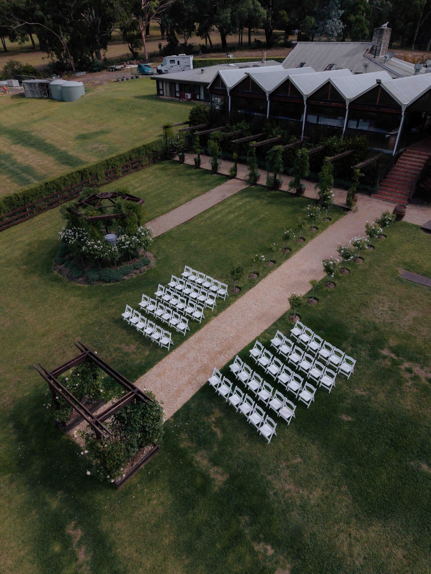 Aerial drone view of outdoor wedding ceremony setup with white chairs on green lawn beside rustic venue building.