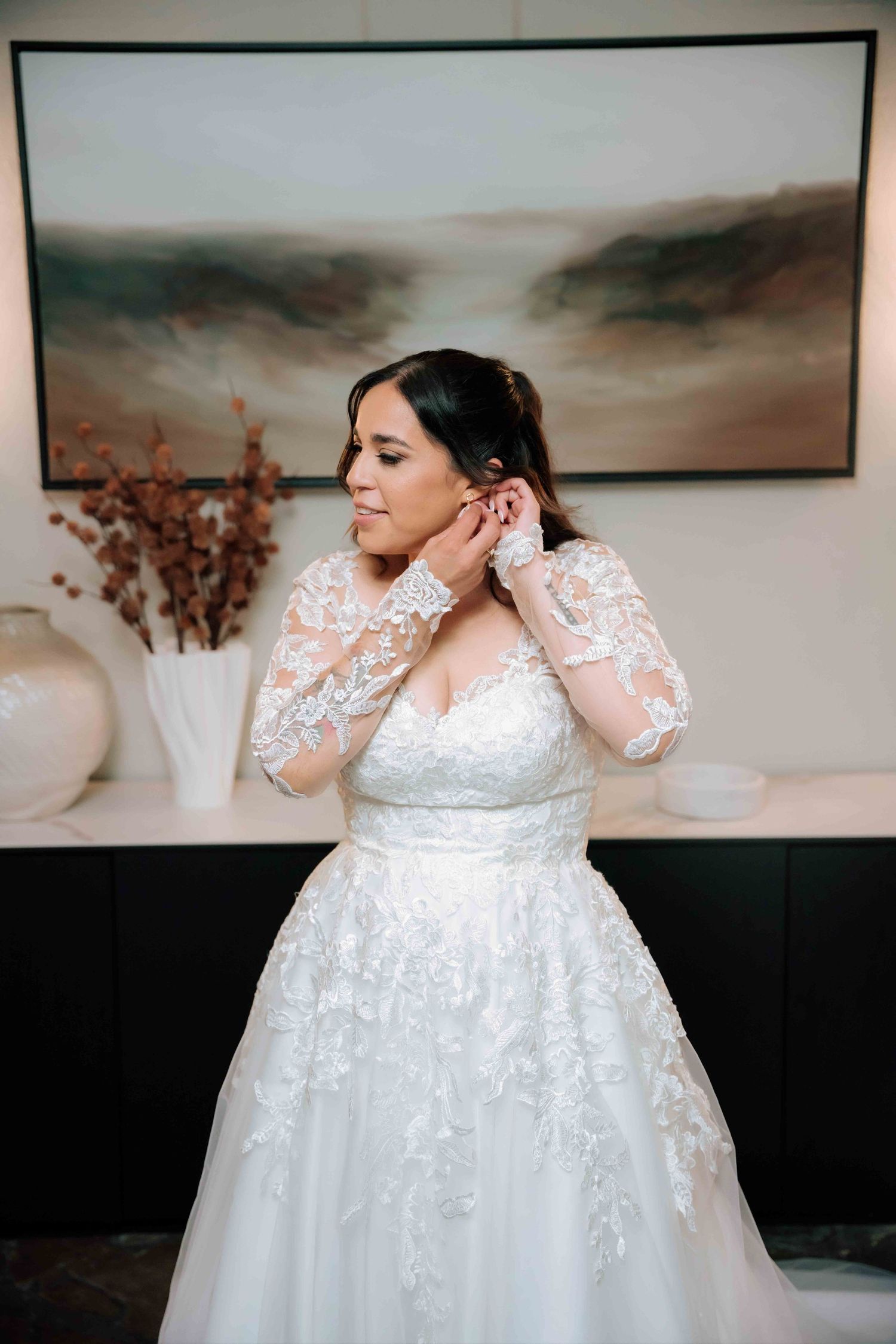 Bride in elegant lace long-sleeve ball gown adjusting earring before floral wall art backdrop.