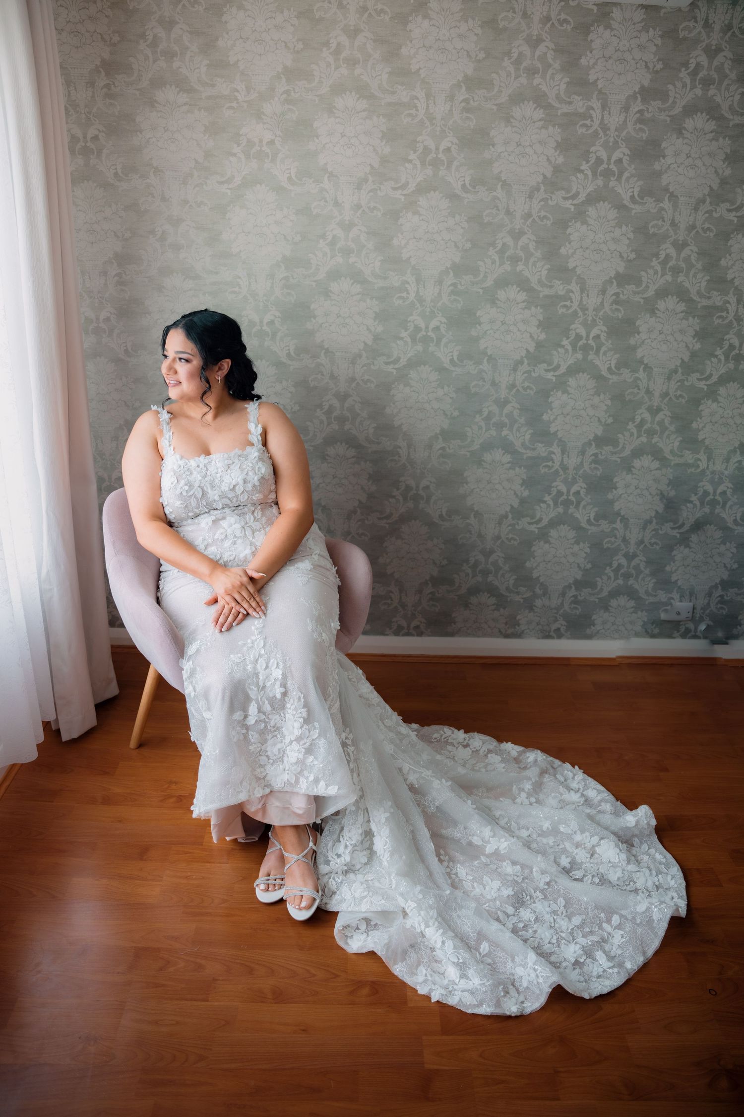 Bride in lace mermaid wedding gown seated in pink chair against elegant damask wallpaper.