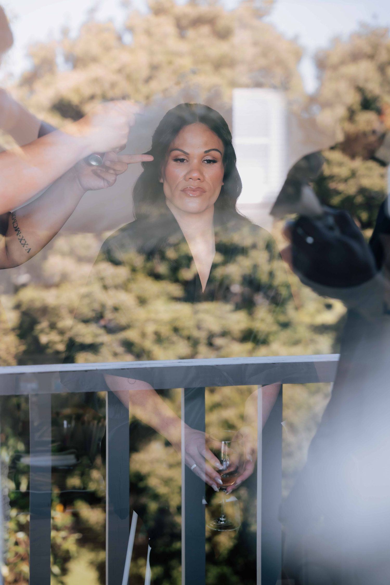 A bridesmaid stands on a balcony holding champagne while someone points at her, with trees in background.