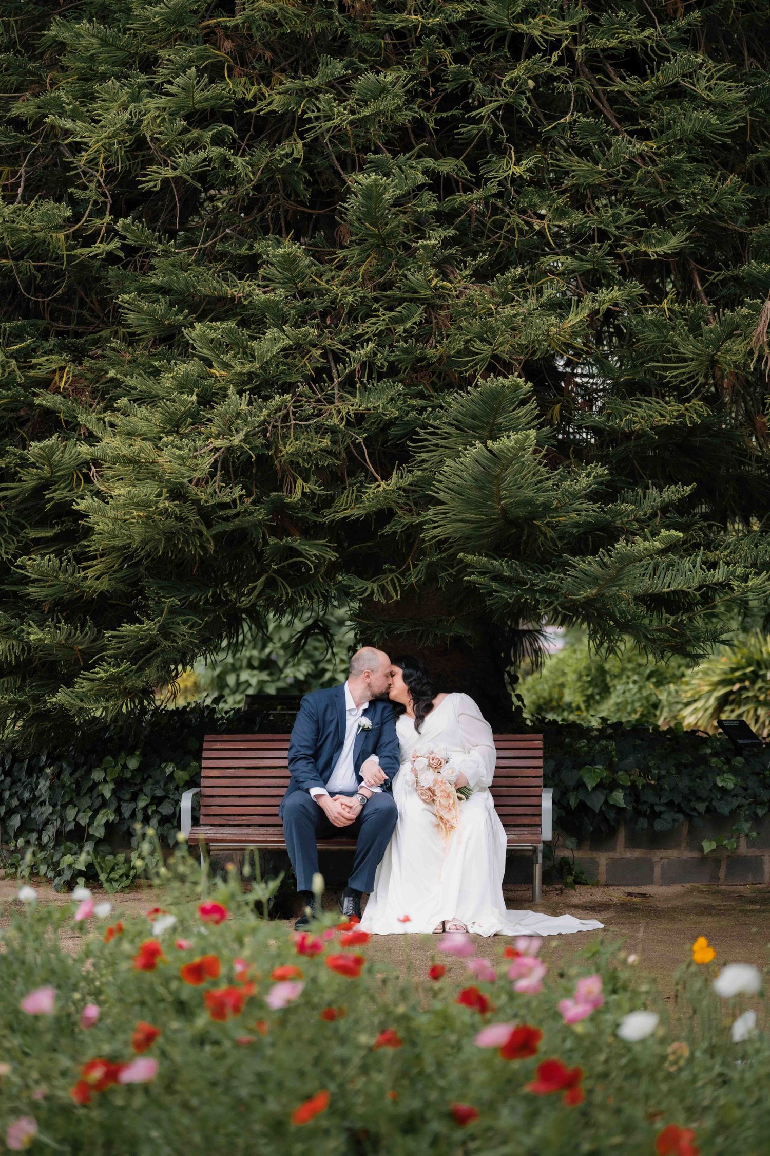 Wedding couple share an intimate moment sitting on a bench surrounded by colorful flowers and evergreen trees.