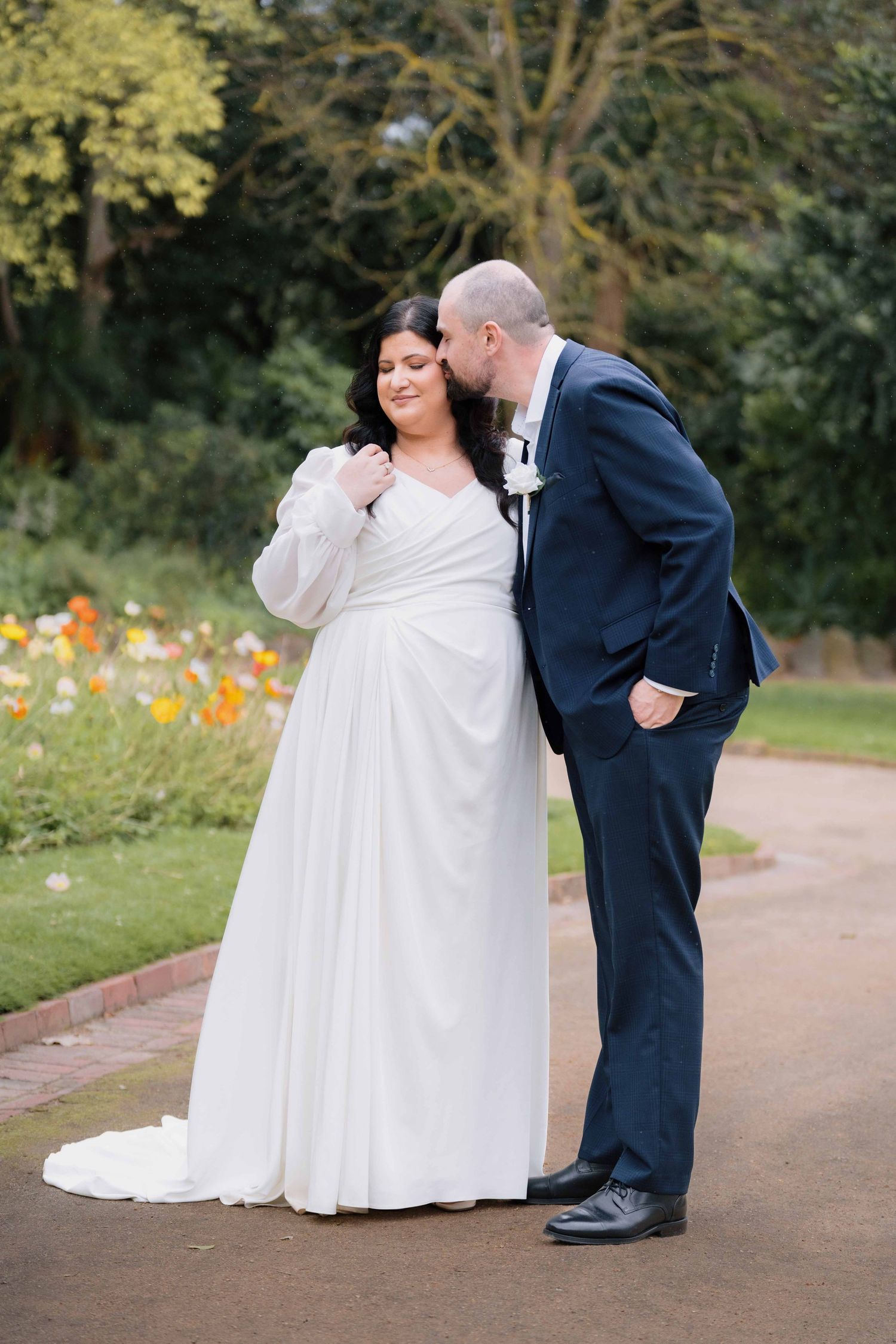 Groom kissing bride on the cheek while she smiles on a garden pathway with flowers.