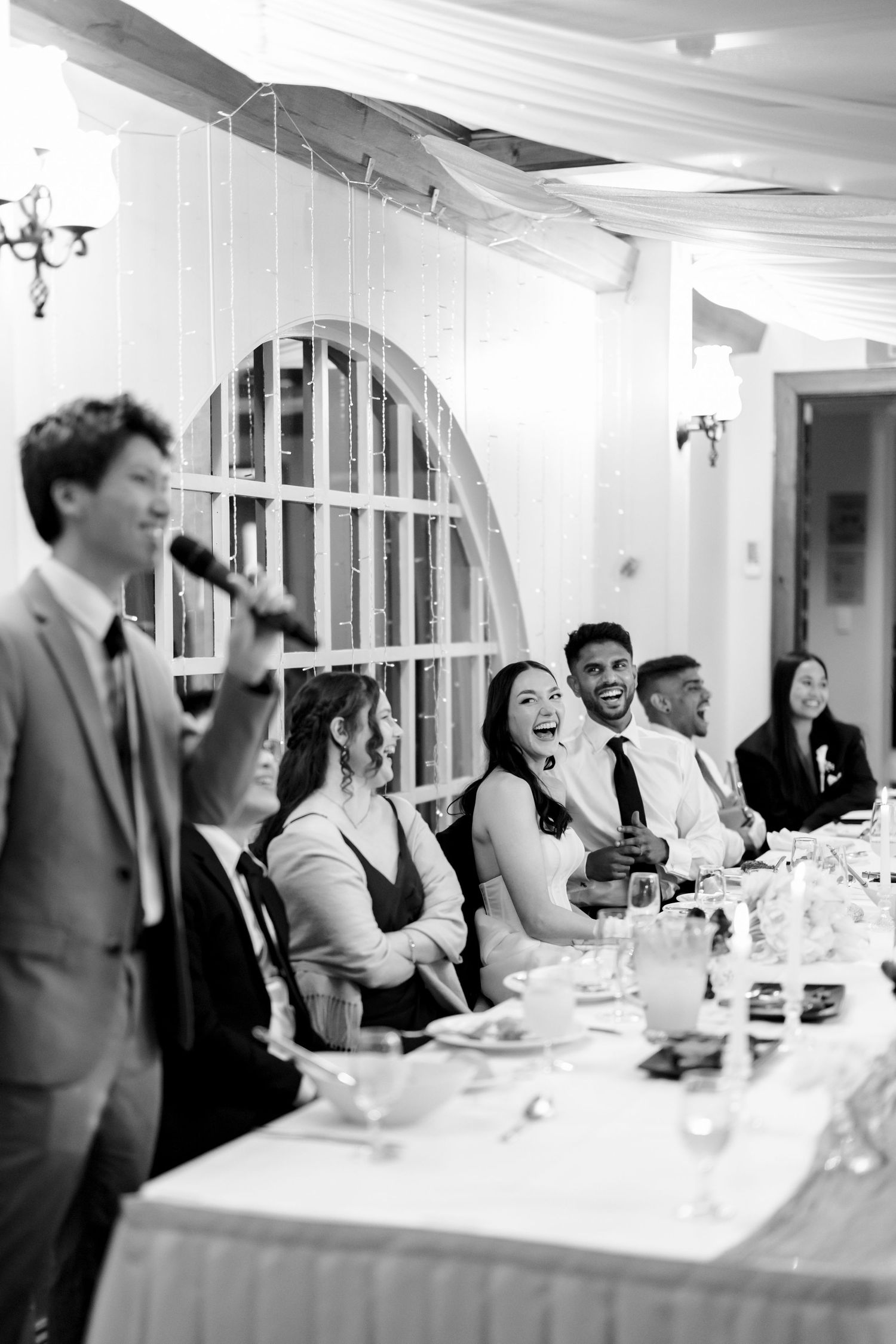 Black and white photo of wedding guests laughing during a toast at an elegant reception dinner.
