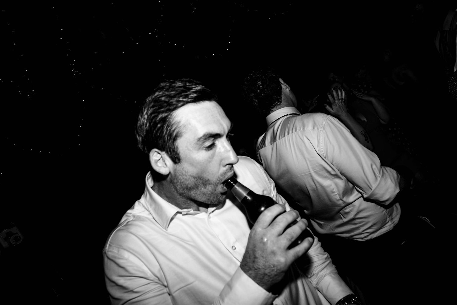 Black and white photo of wedding guest drinking from a bottle on dance floor.
