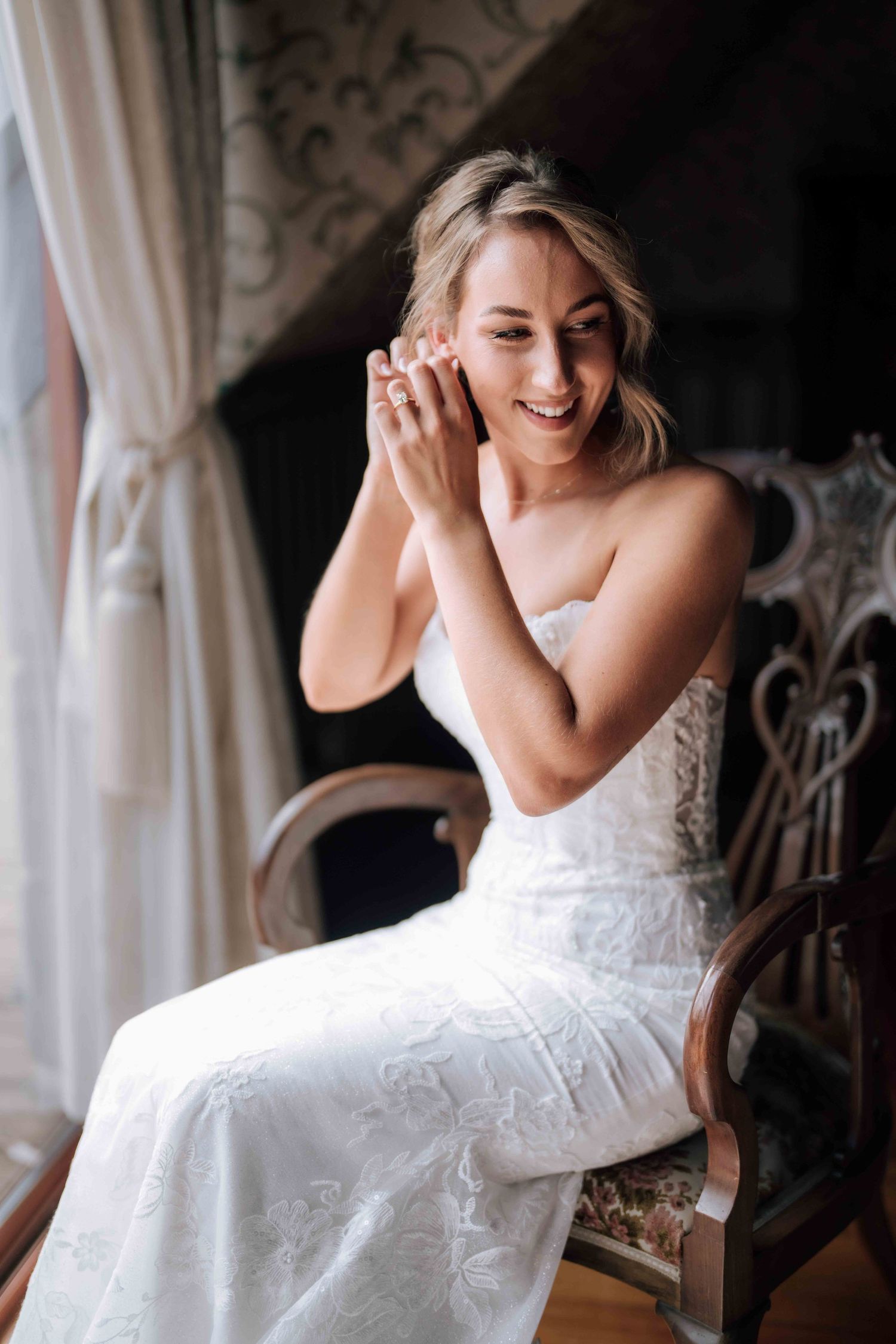 Smiling bride in strapless white lace wedding dress adjusting earring while seated on ornate chair.