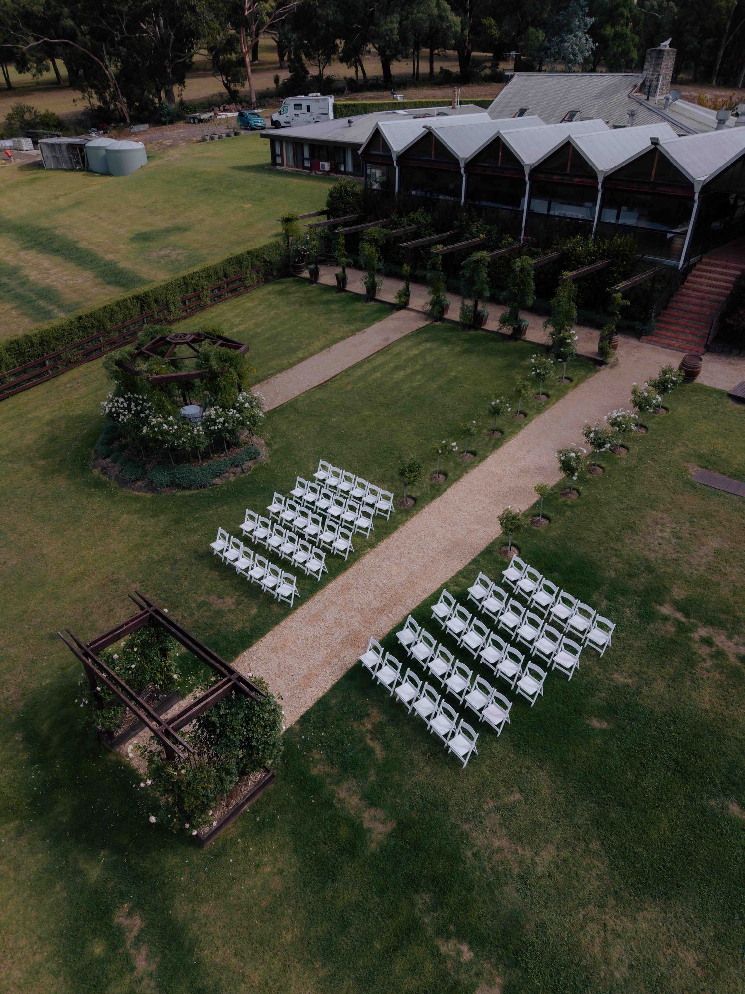 Aerial view of outdoor wedding ceremony setup with white chairs along a garden path beside a rustic venue.