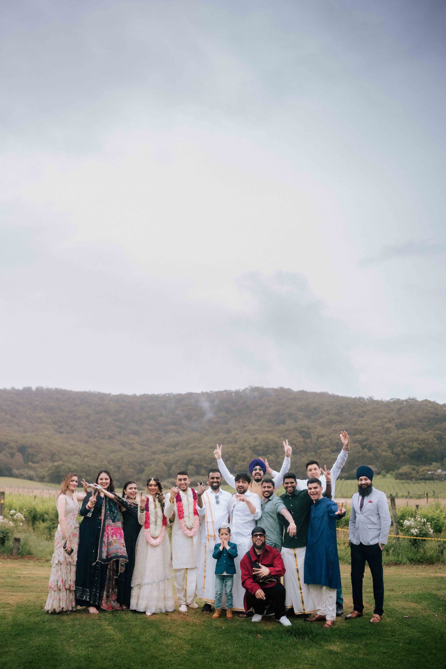 Indian wedding group photo with guests in traditional attire posing on green lawn with mountain backdrop.