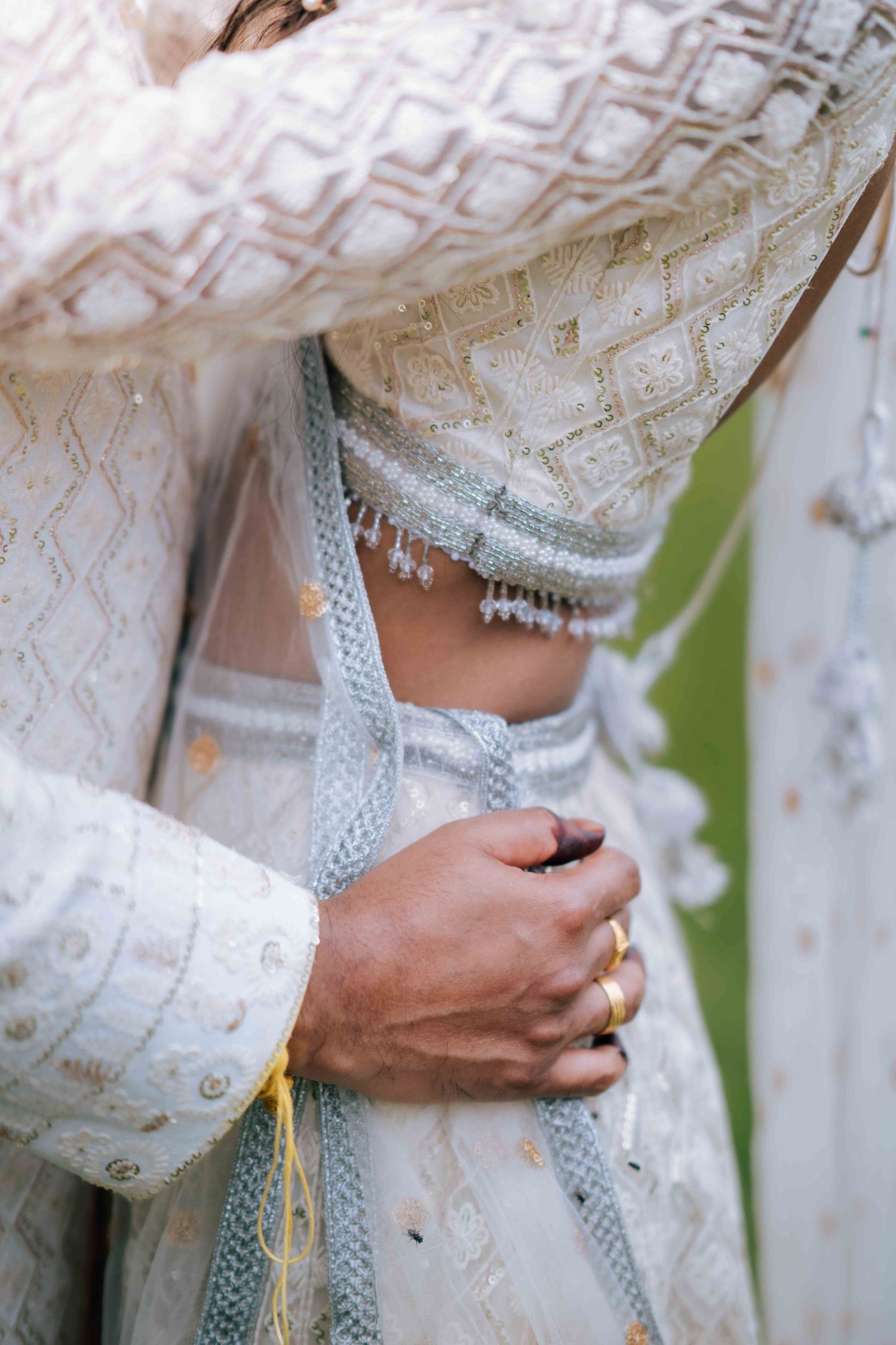 Close-up of Indian bride in white lehenga with silver waist jewelry and groom's hand with gold ring.