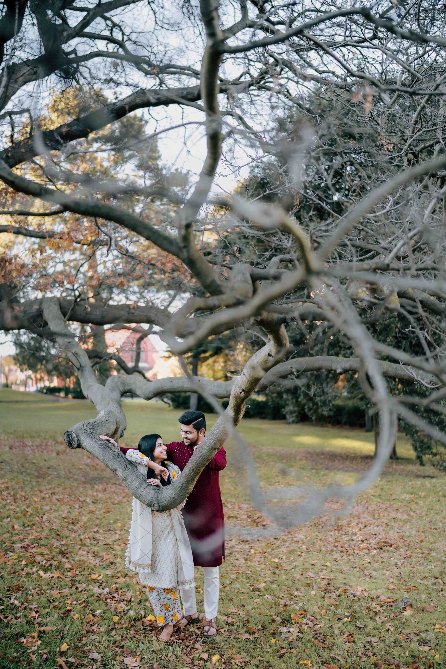 Couple in traditional Indian outfits sharing a tender moment leaning against a curved bare tree branch.