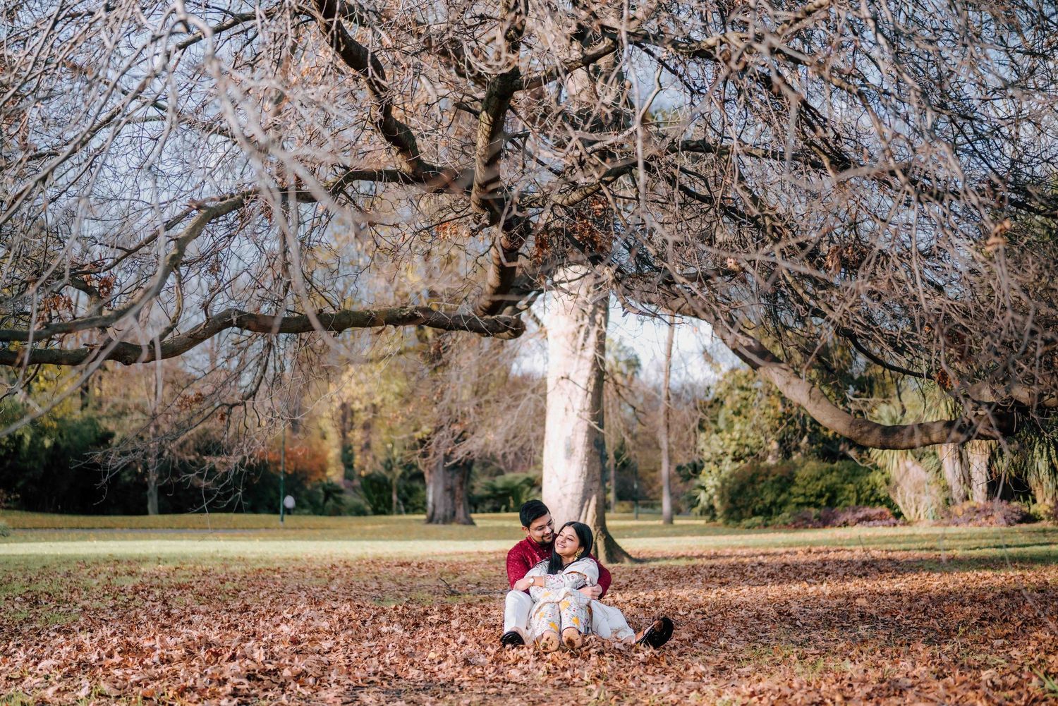 Couple relaxing together among fallen autumn leaves beneath a bare tree in a peaceful park setting.