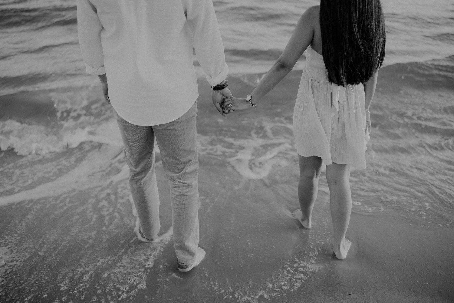 Black and white photo of couple holding hands while walking in ocean waves on a beach.