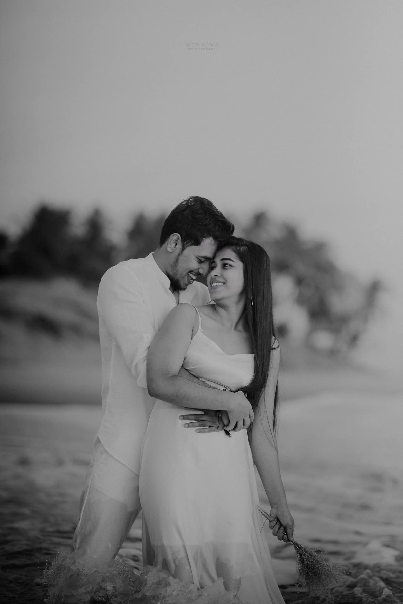Black and white romantic beach photo of couple embracing with palm trees and ocean in background.