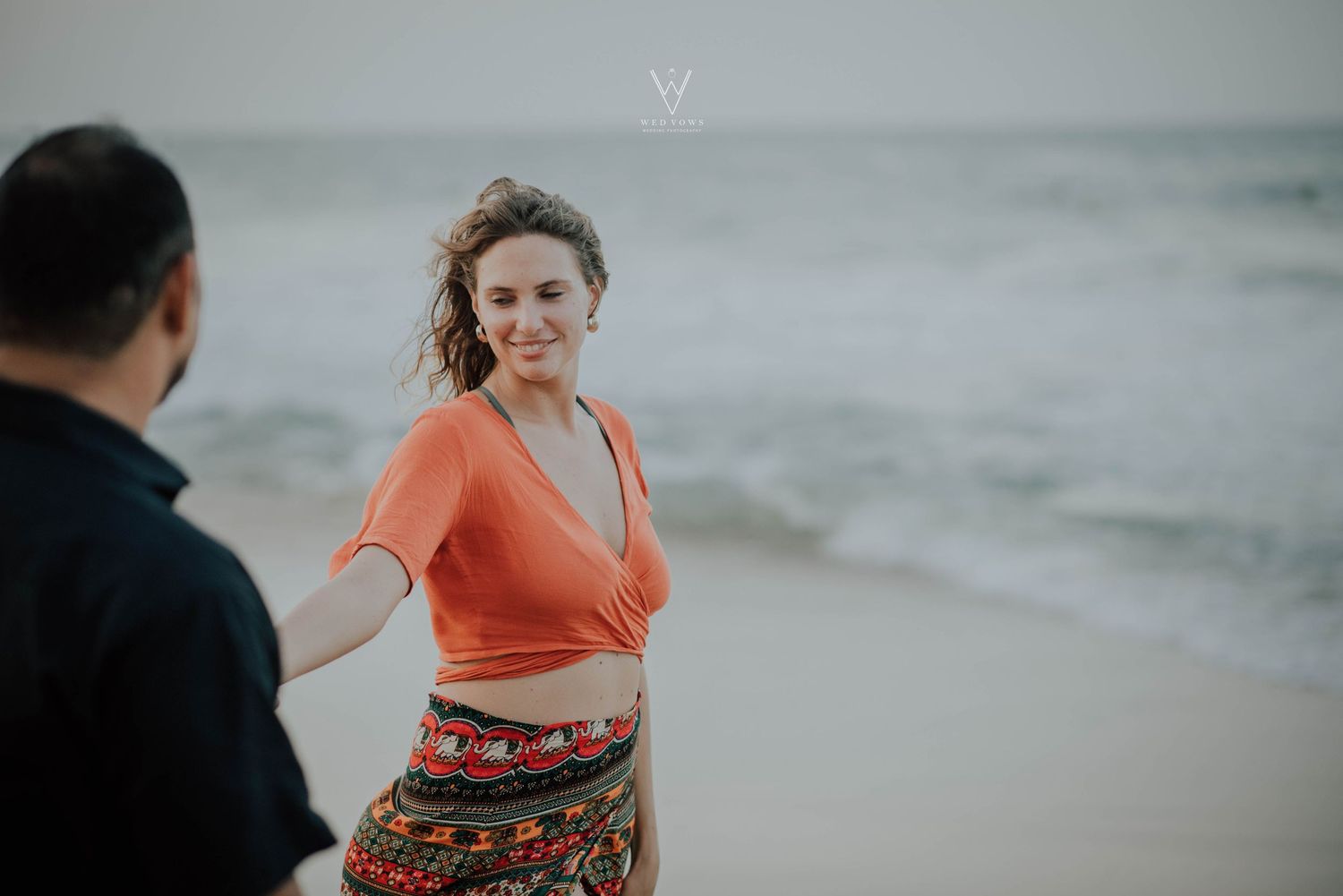 Woman in orange top smiling while holding hands with partner on a beach.