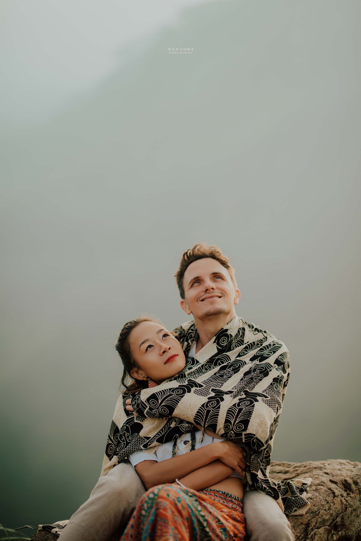 Couple wrapped in batik fabric sitting together gazing upward in misty mountain setting.