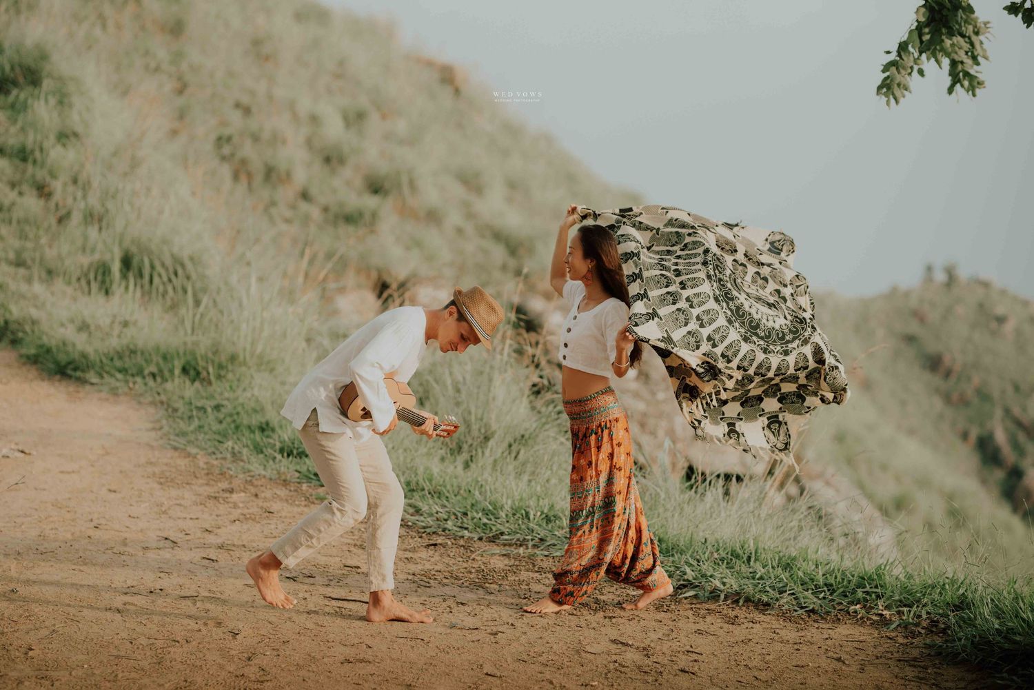 A playful couple walks along a dirt hillside path, woman carrying a large mandala blanket overhead in warm golden light.