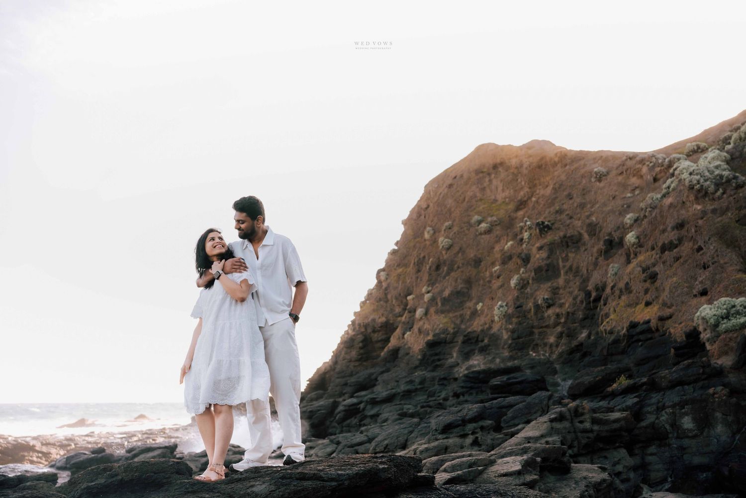 Couple in matching white outfits embracing on rugged coastal rocks with ocean in background.