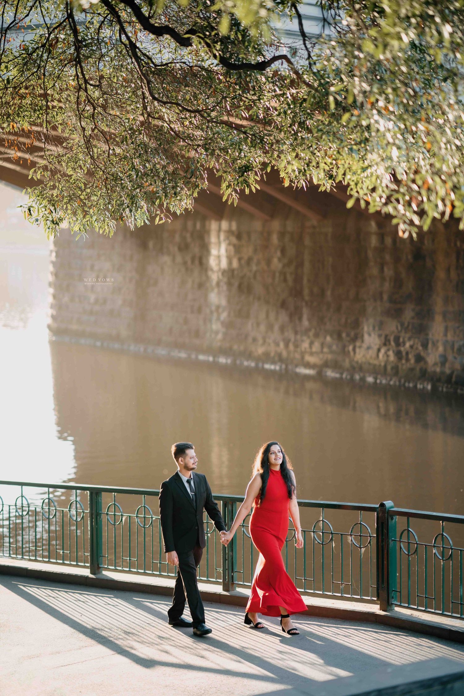 Couple holding hands walk along a sunlit riverside promenade, woman in elegant red halter dress.